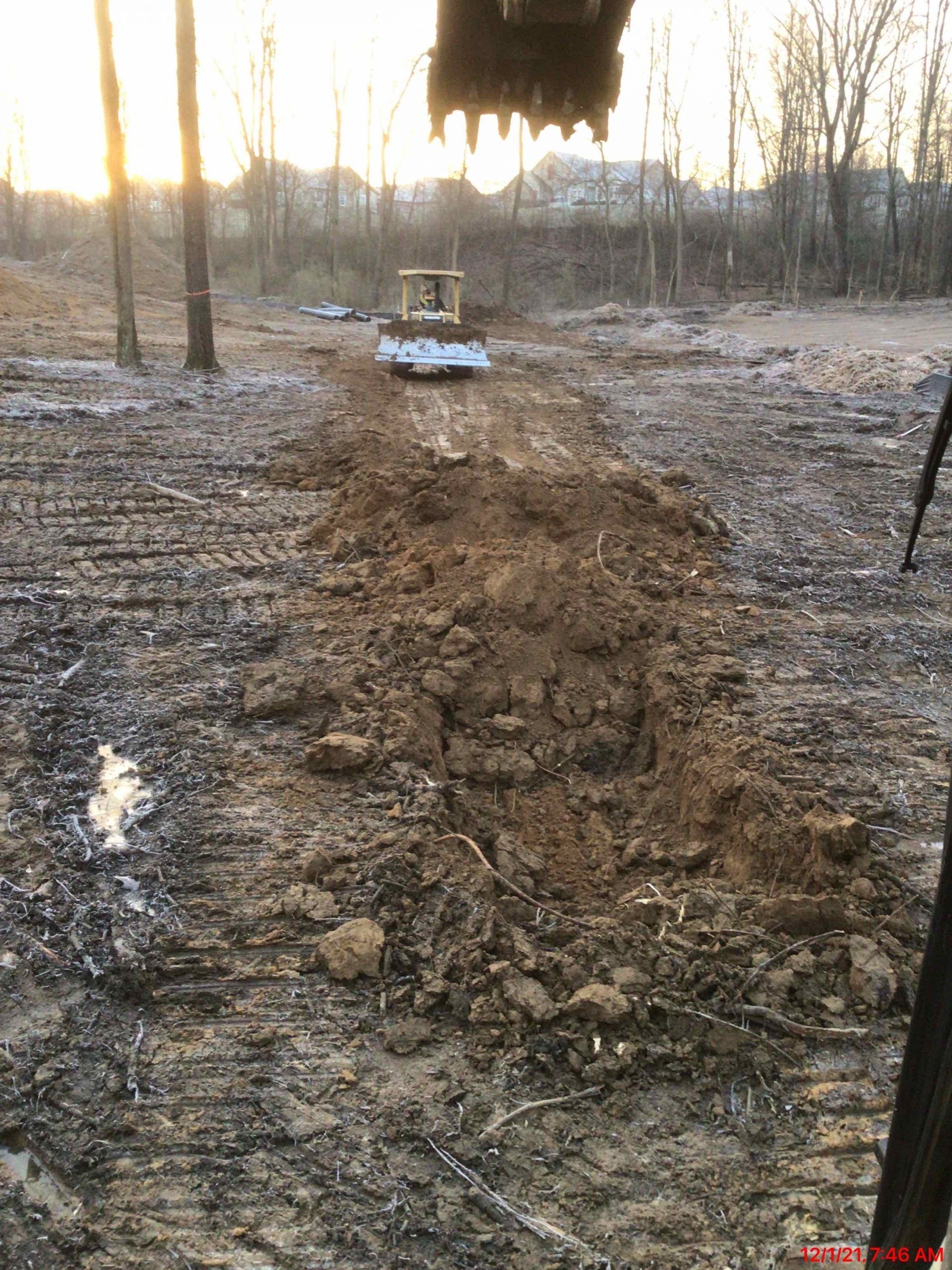 An excavator digging a trench in muddy ground. Setting is outdoors with trees and a box in the distance.