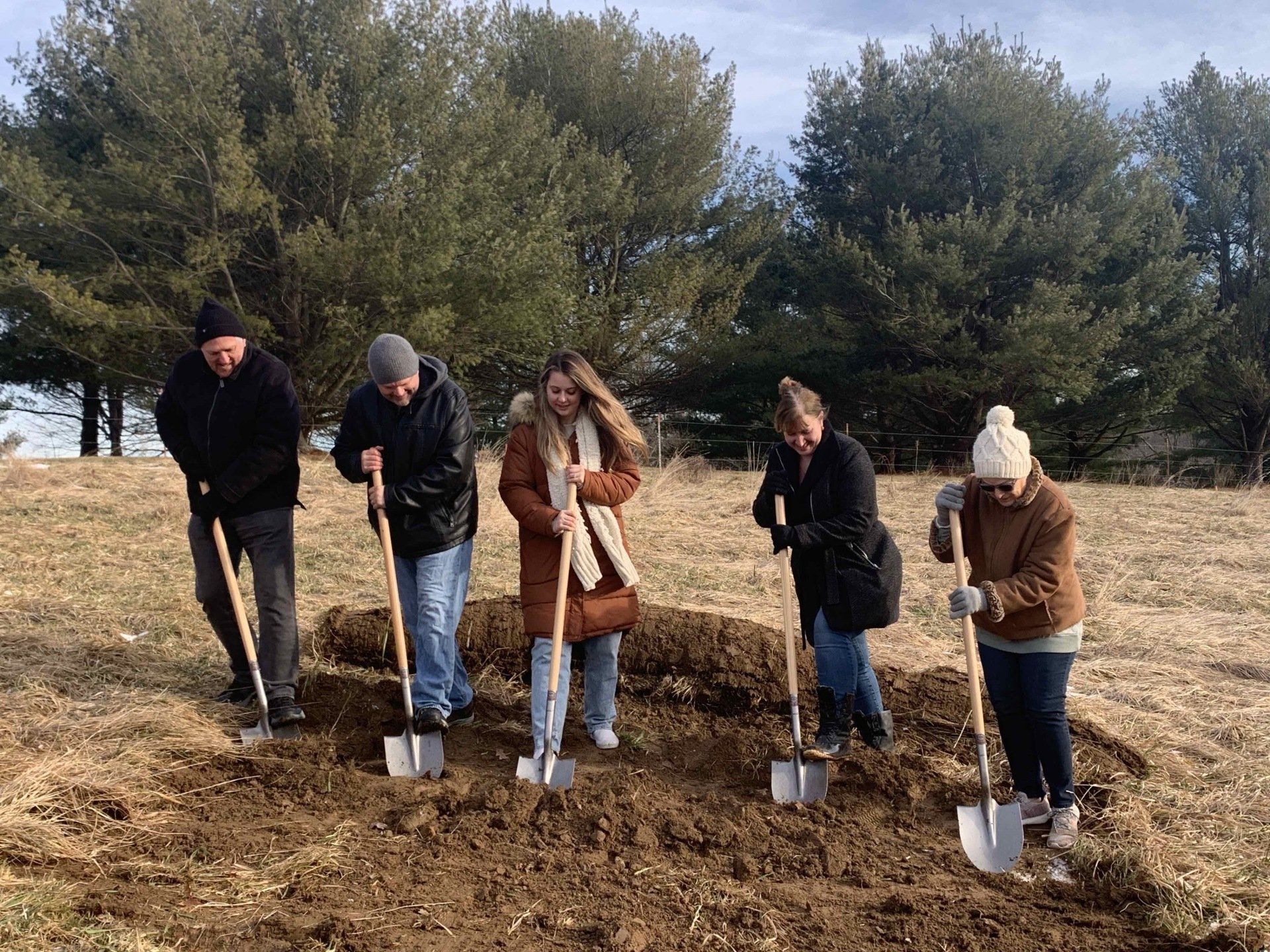 Five people shoveling dirt on a field with trees in the background, possibly a groundbreaking ceremony.