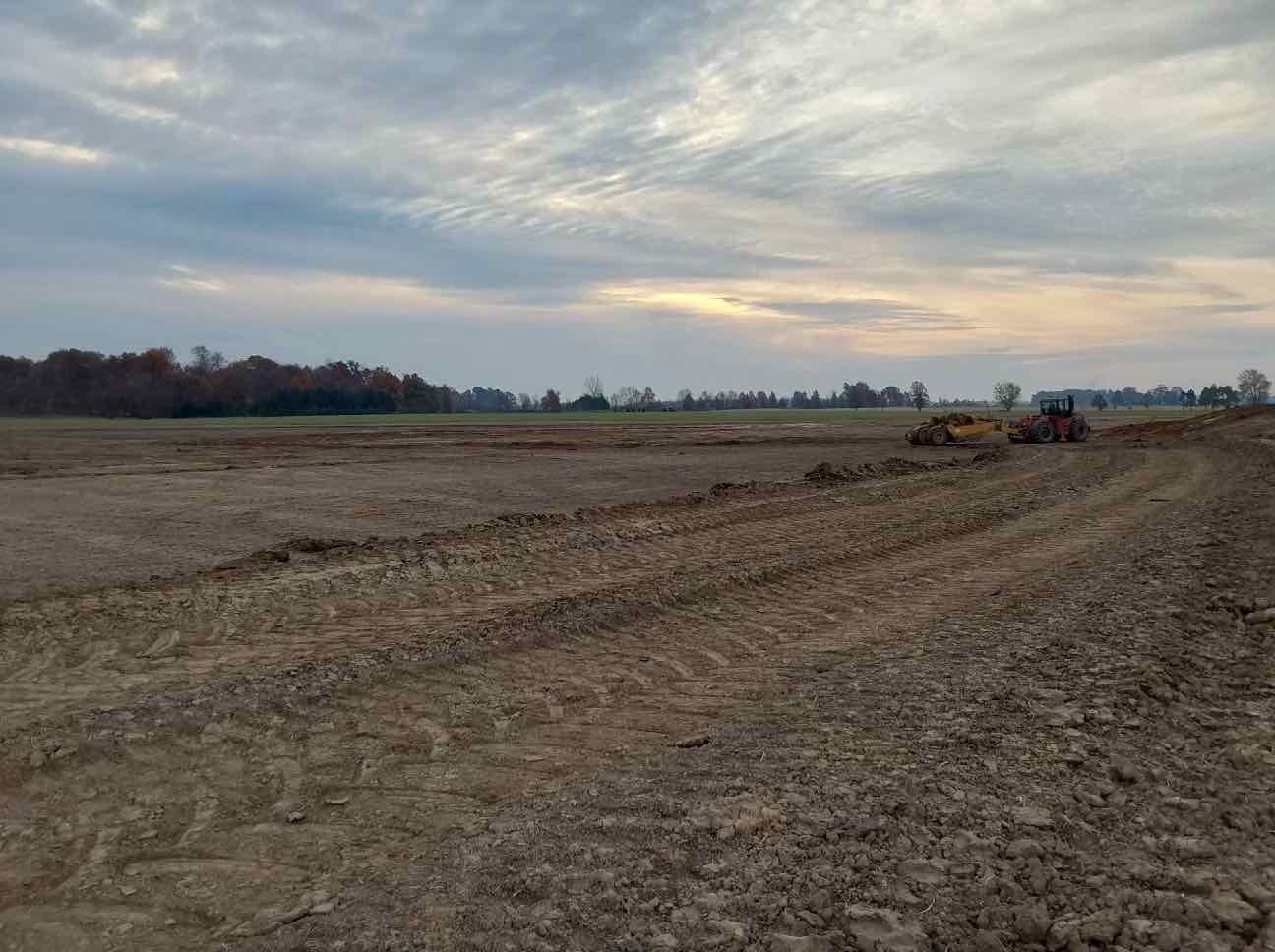 A freshly tilled brown field under a cloudy sky. A yellow construction vehicle is visible in the distance.