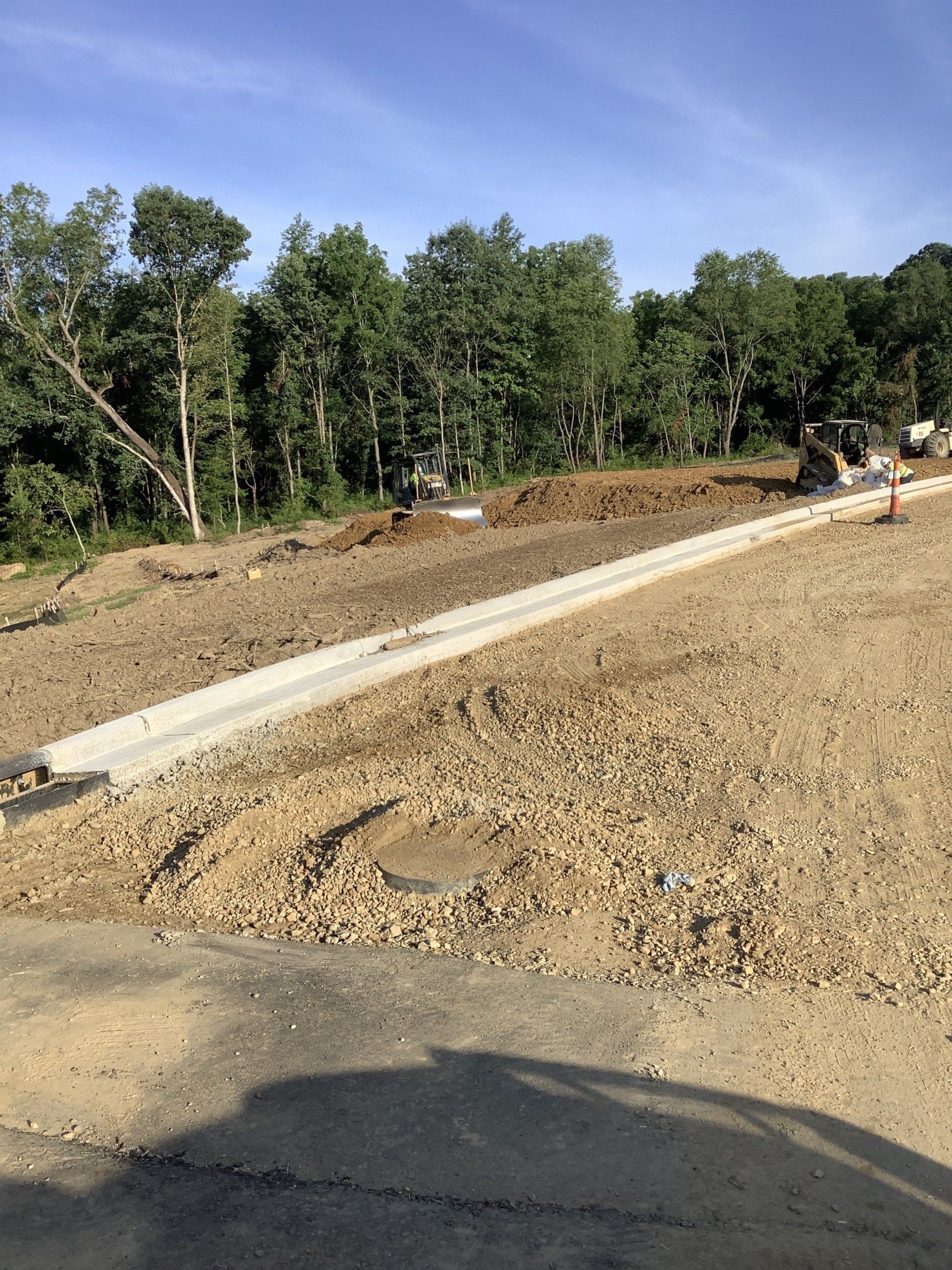 Construction site with concrete curb and gravel, trees in background, sunny day.
