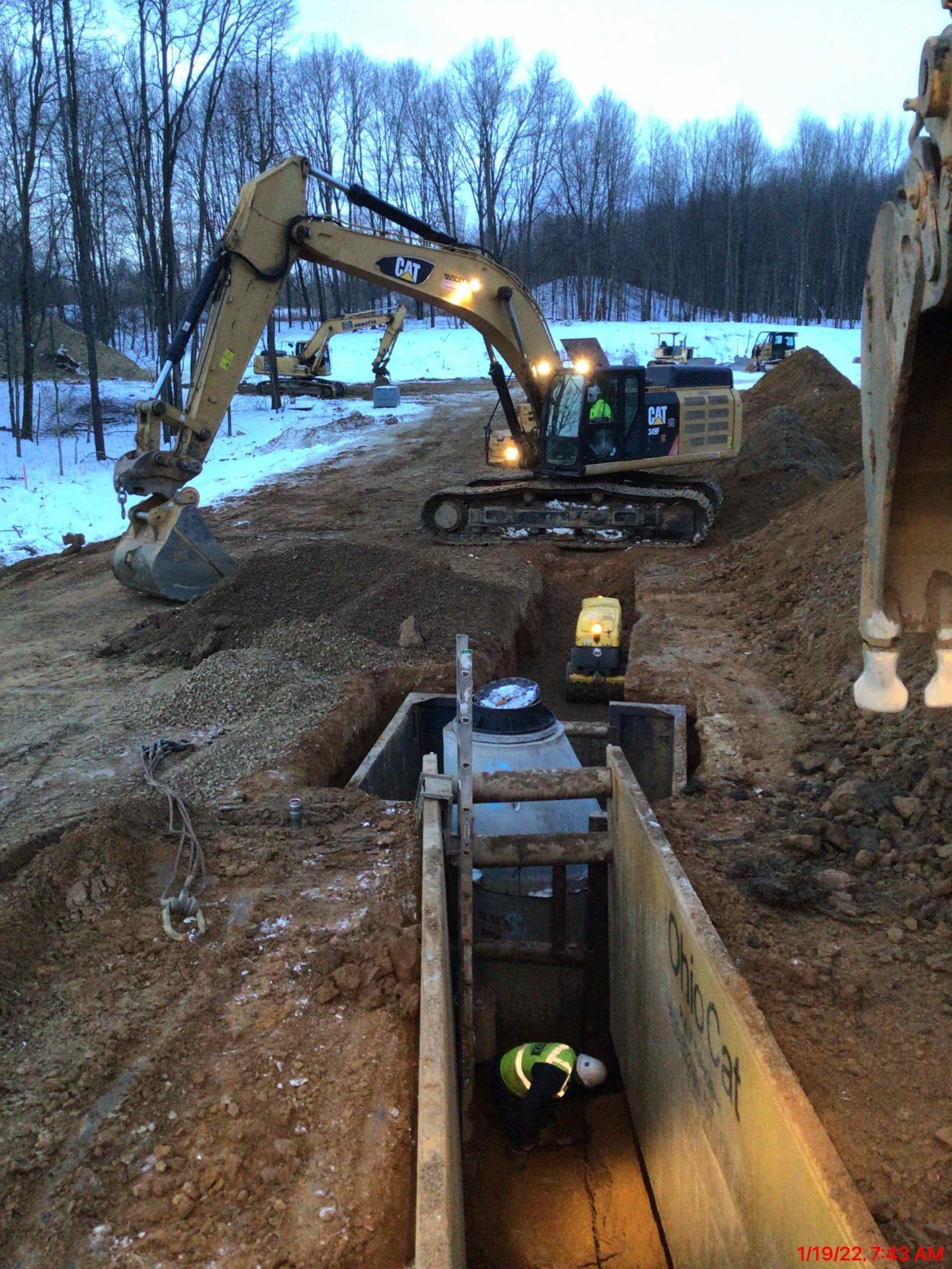 An excavator and worker in a trench. The worker is wearing a safety vest. Construction site.