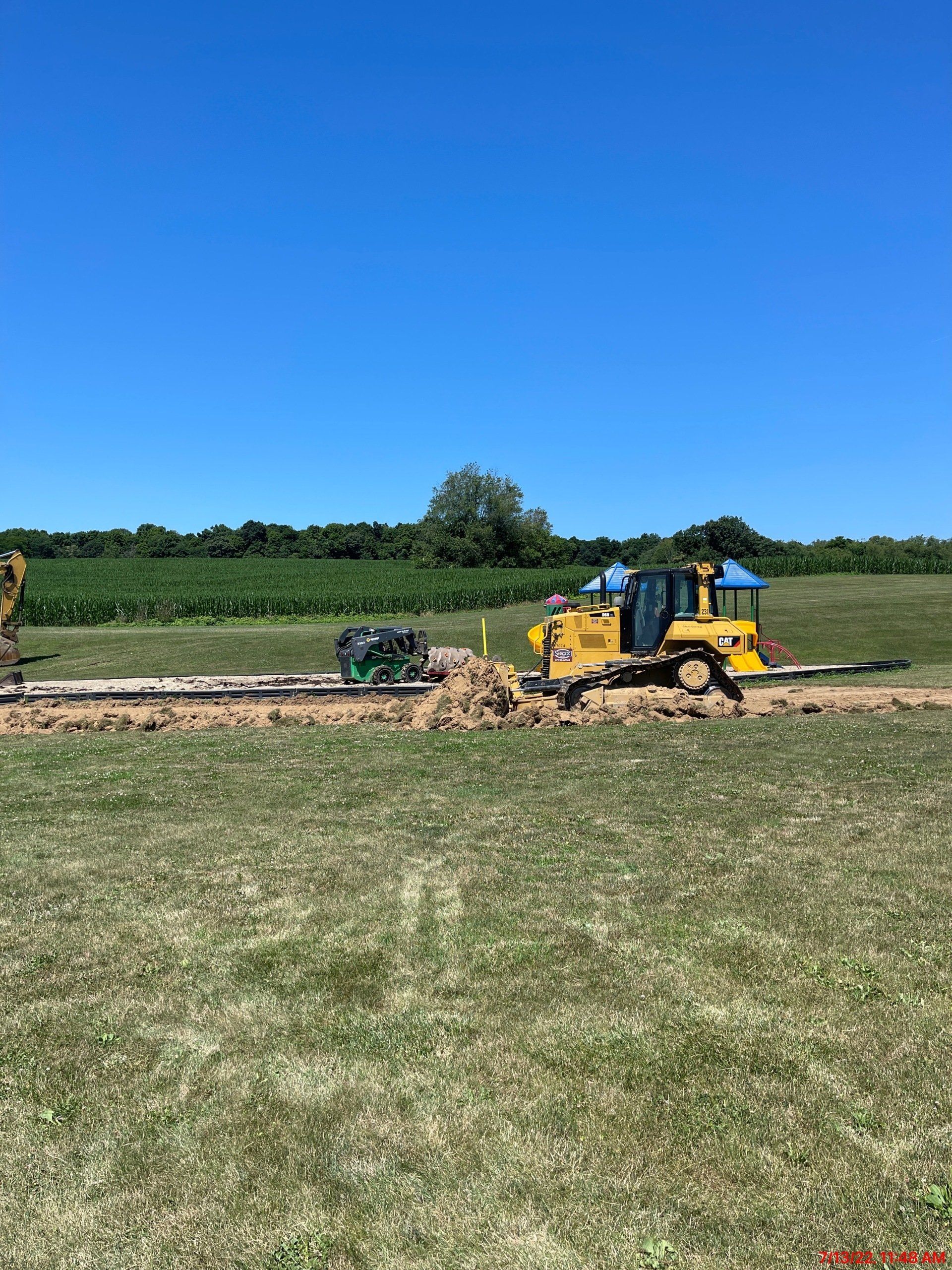 Yellow bulldozer burying a pipeline in a grassy field under a blue sky.