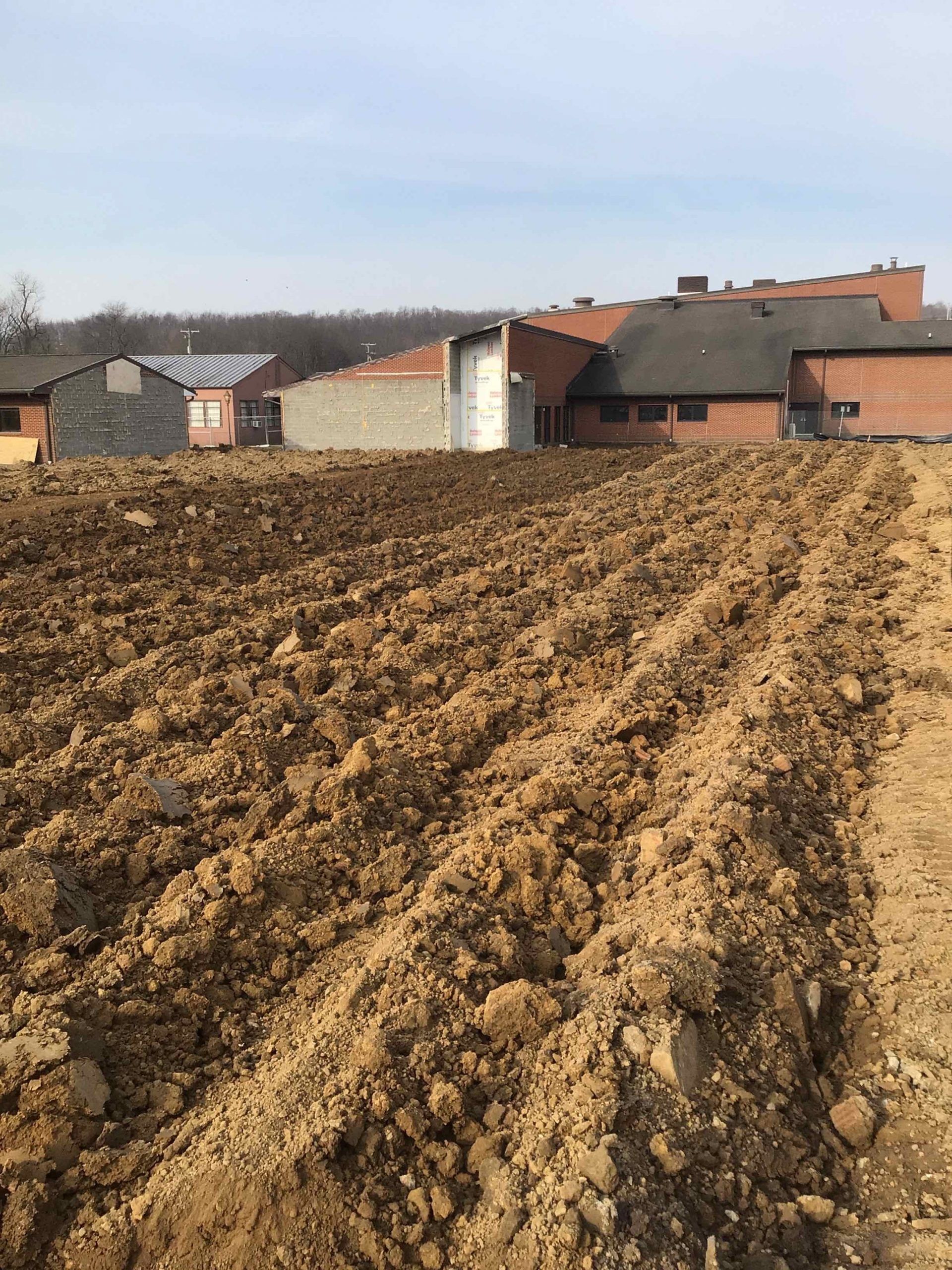 Plowed brown field in the foreground; buildings with red brick and gray walls in the background under a blue sky.