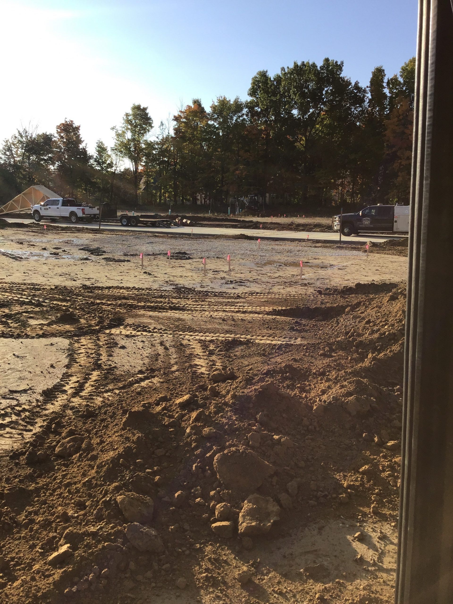 Construction site with dirt, tire tracks, vehicles, and trees in background.