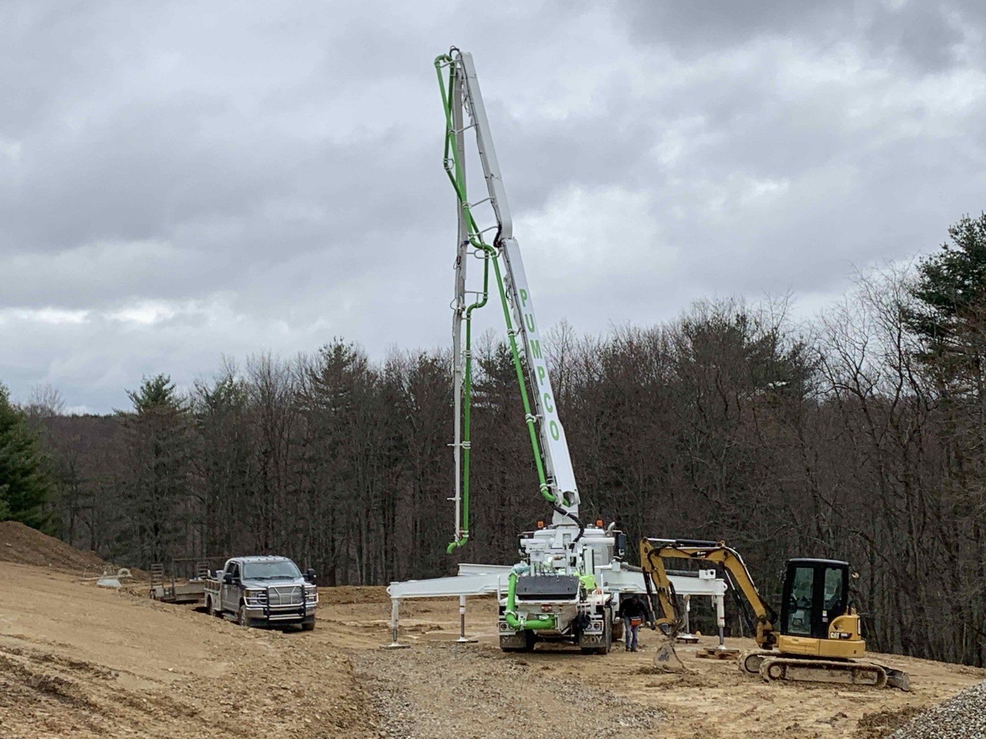 Concrete pump truck on construction site, boom extended. Cloudy sky, trees in the background.