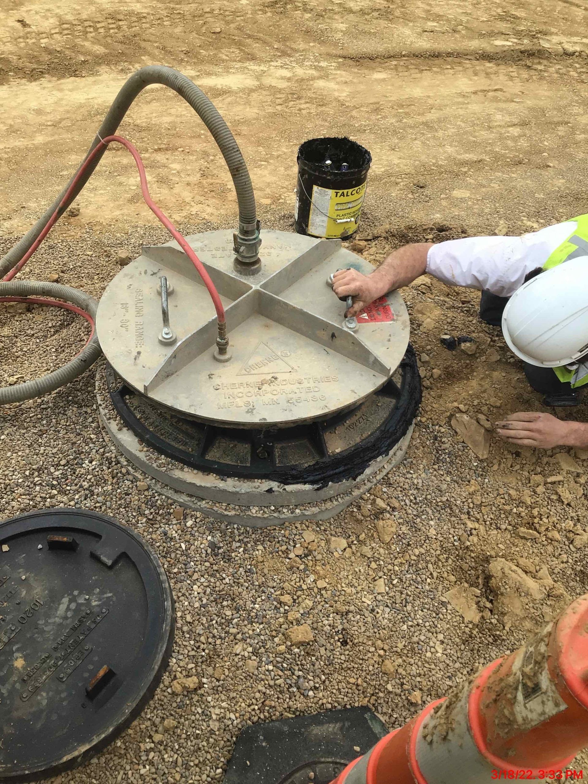 Worker in hard hat repairs utility access cover with attached hose. The cover is on gravel; a bucket sits nearby.