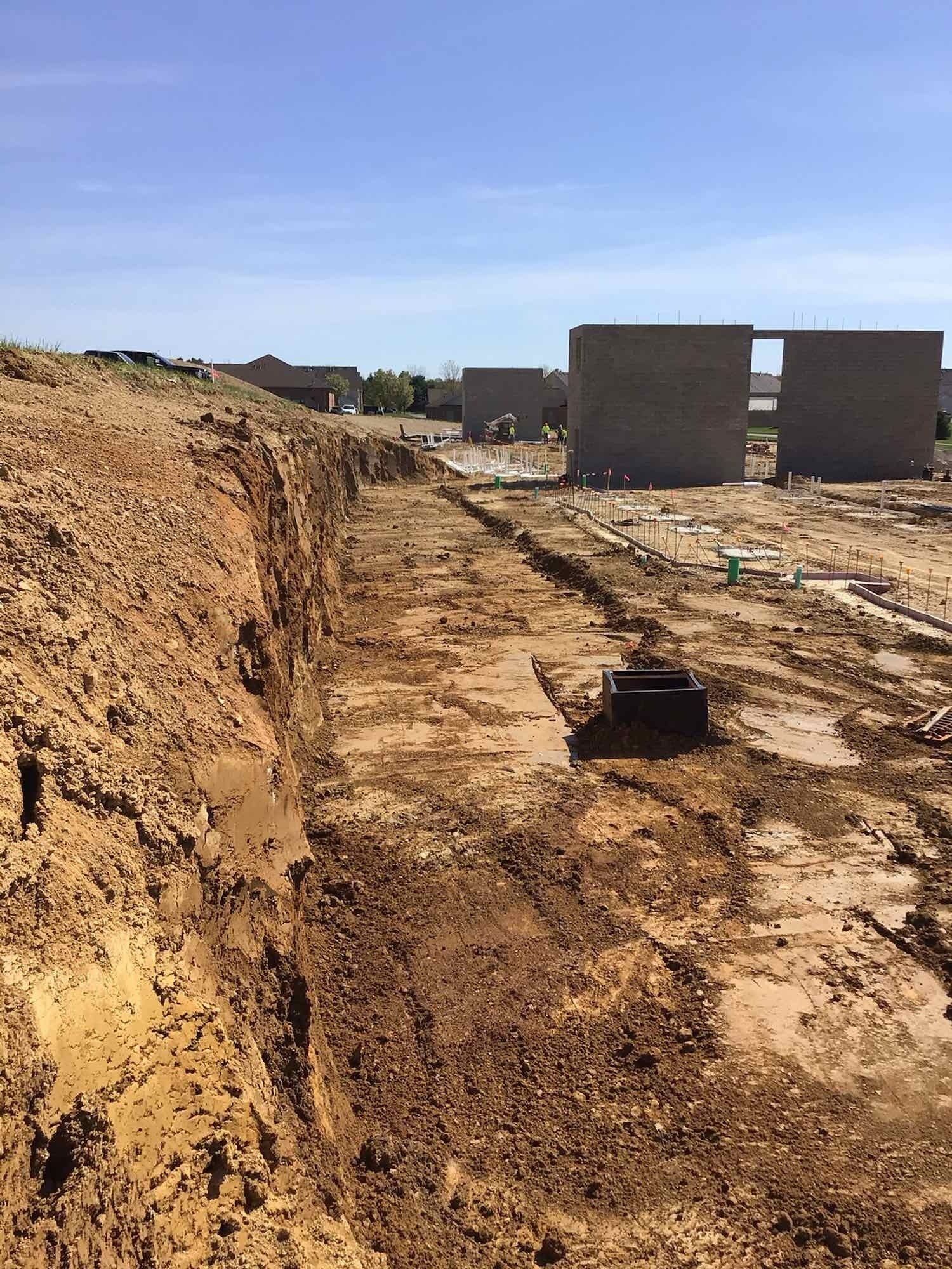 Construction site with trenches in muddy ground. Buildings under construction in the distance.