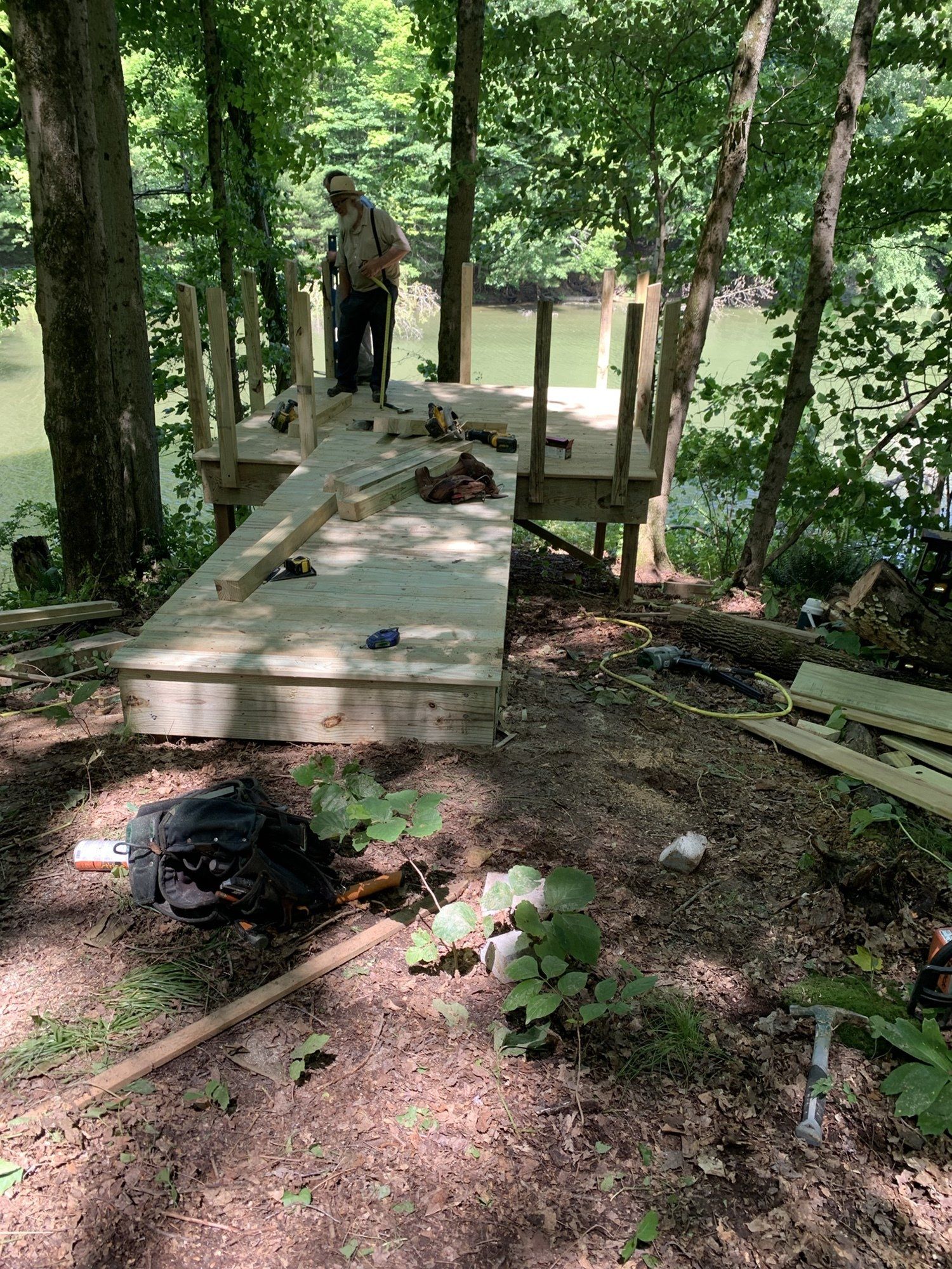 A person standing on a newly constructed wooden deck with a ramp in a wooded area near water.