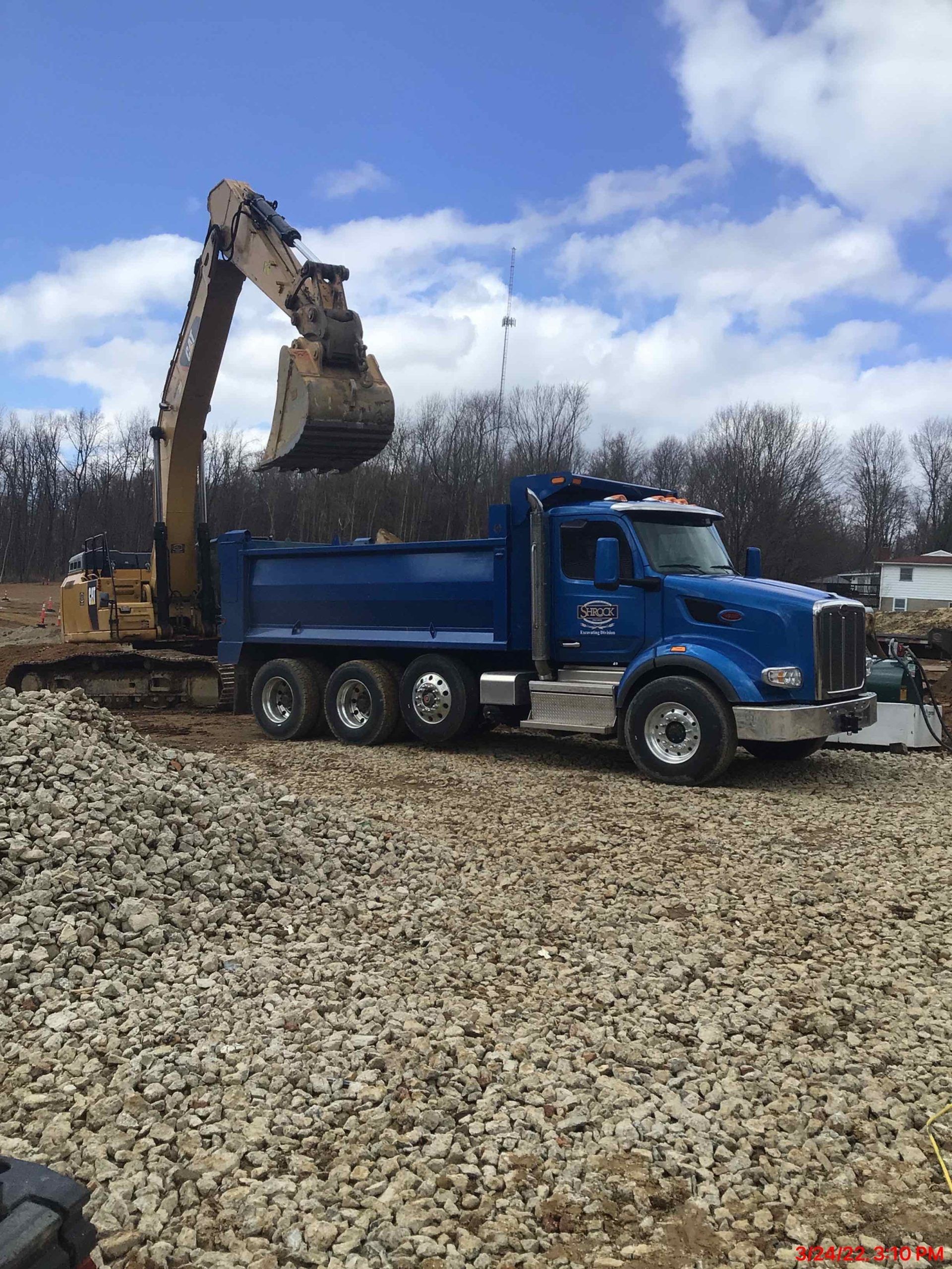 Blue dump truck being loaded by a yellow excavator at a construction site on a cloudy day.