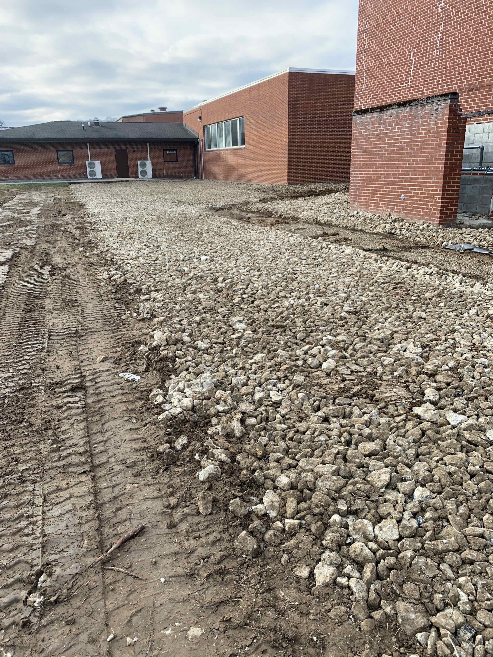 Construction site with brick buildings and gravel path, muddy tire tracks. Overcast day.