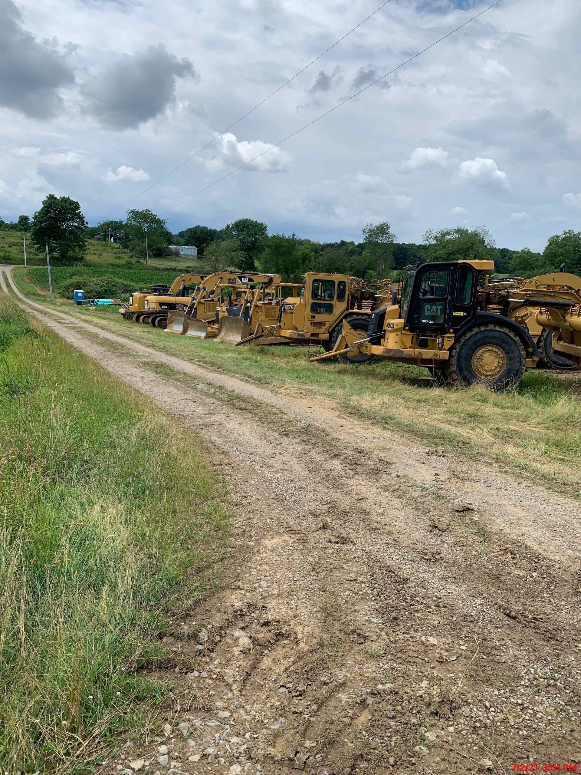 Dirt road lined with yellow construction equipment on a grassy field under cloudy sky.