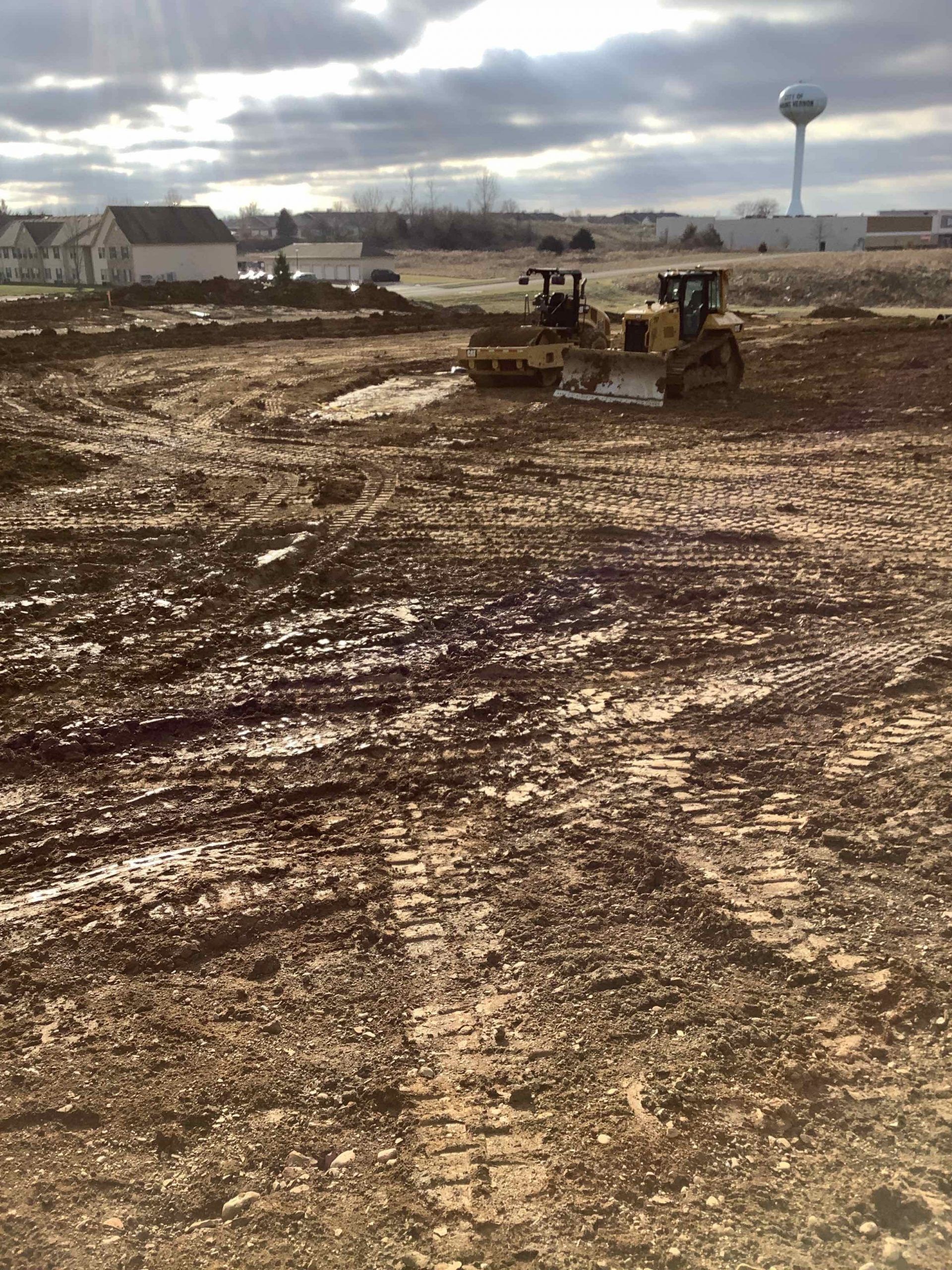 Two bulldozers on muddy construction site, overcast sky, water tower in background.
