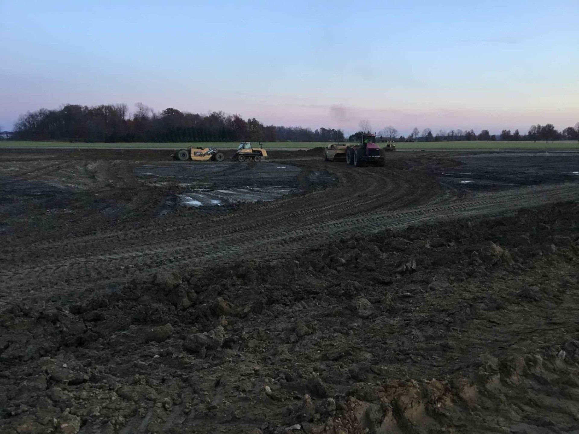 Construction site with heavy machinery on muddy ground, dusk sky, and treeline in the distance.