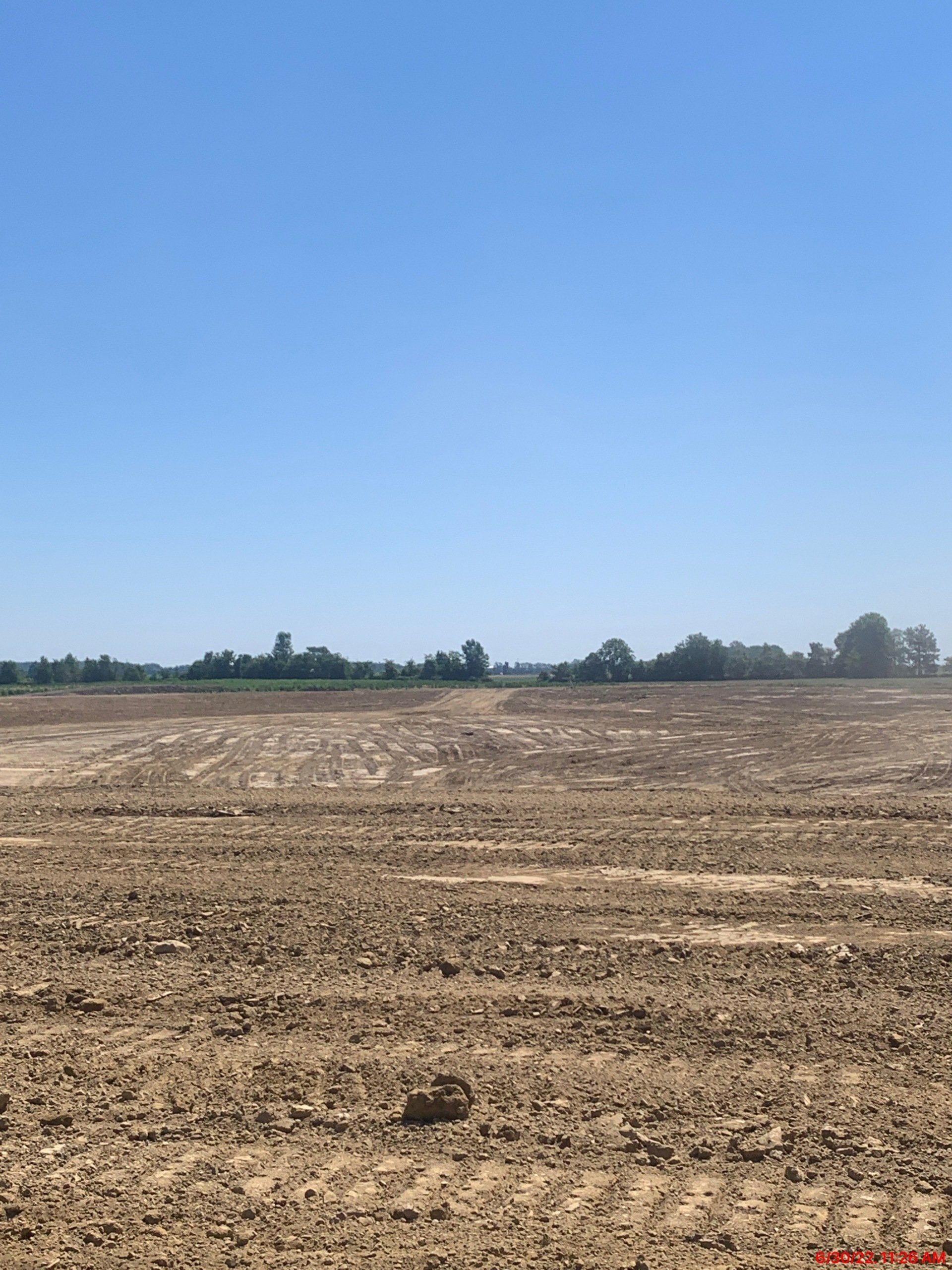 Tilled brown field under a clear blue sky, distant trees and a structure visible.