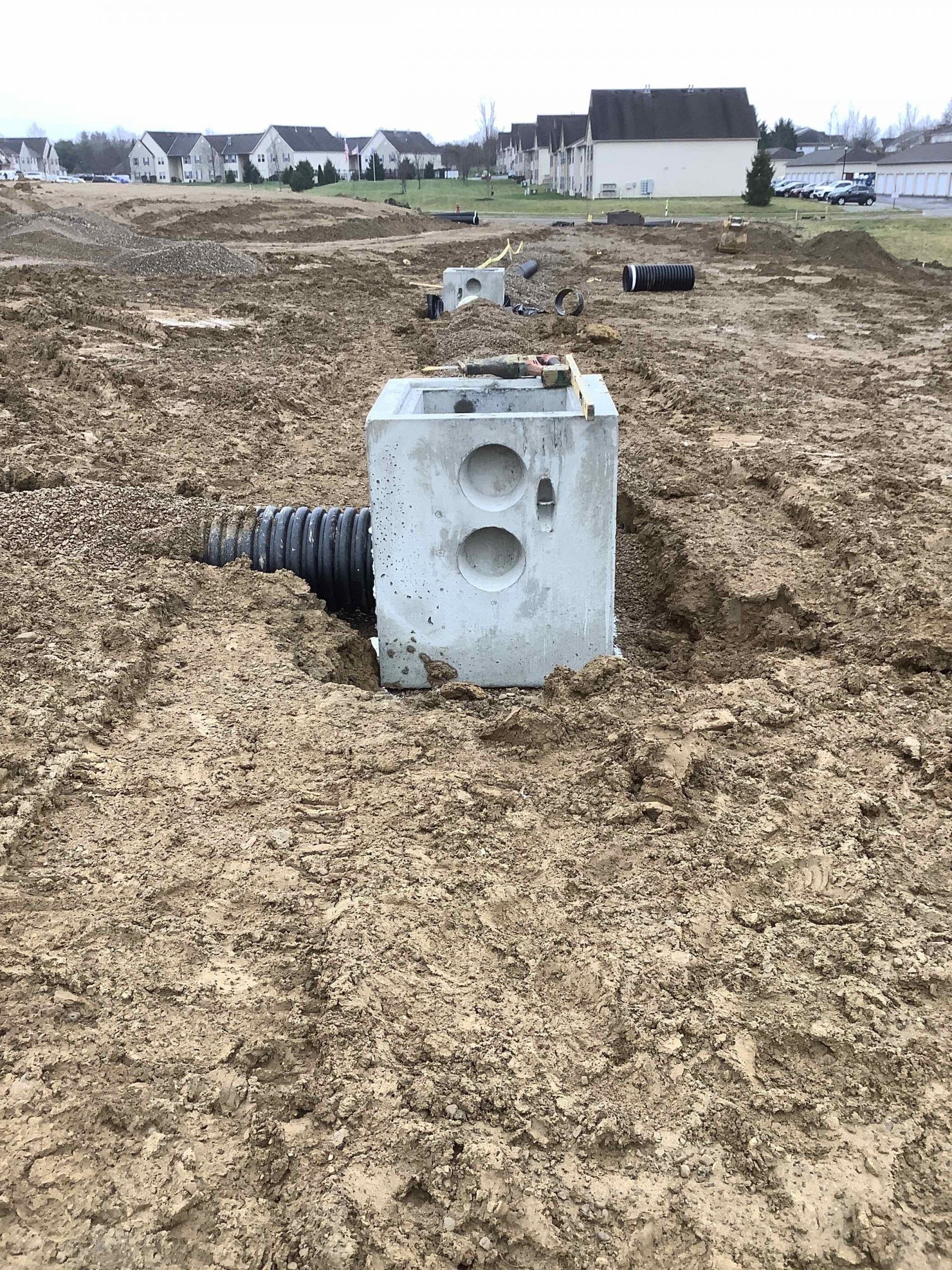 Concrete drainage structure with corrugated pipe in muddy construction site. Houses in background.