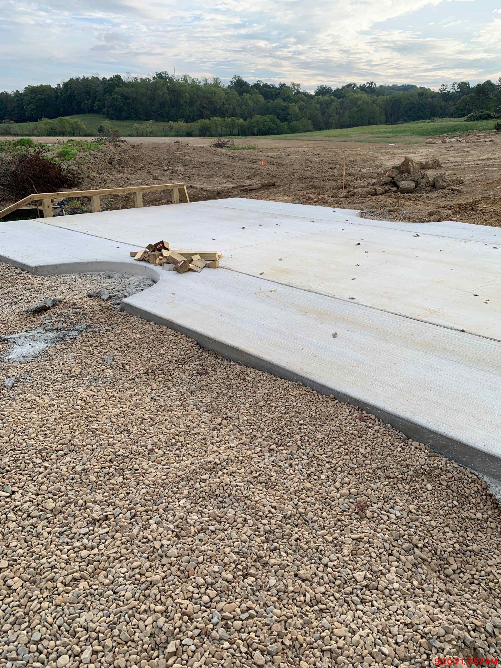 Concrete slab and gravel ground with construction materials, trees in the background.