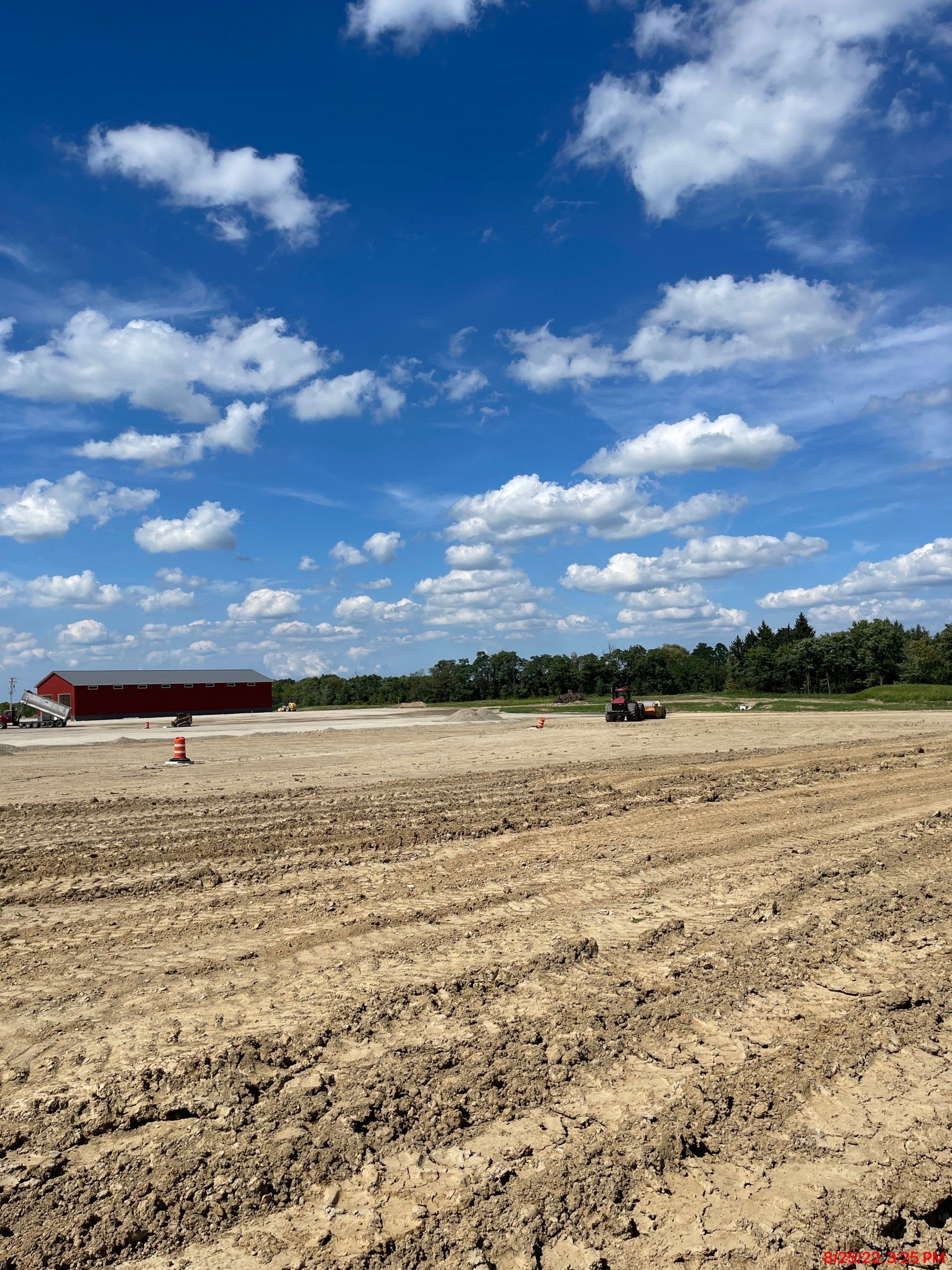 A large, plowed field under a bright blue sky with scattered clouds, with a red barn in the distance.