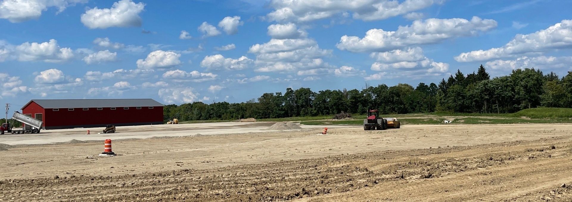 A red barn sits near a construction site with a tractor on a sunny day. Trees in the background.