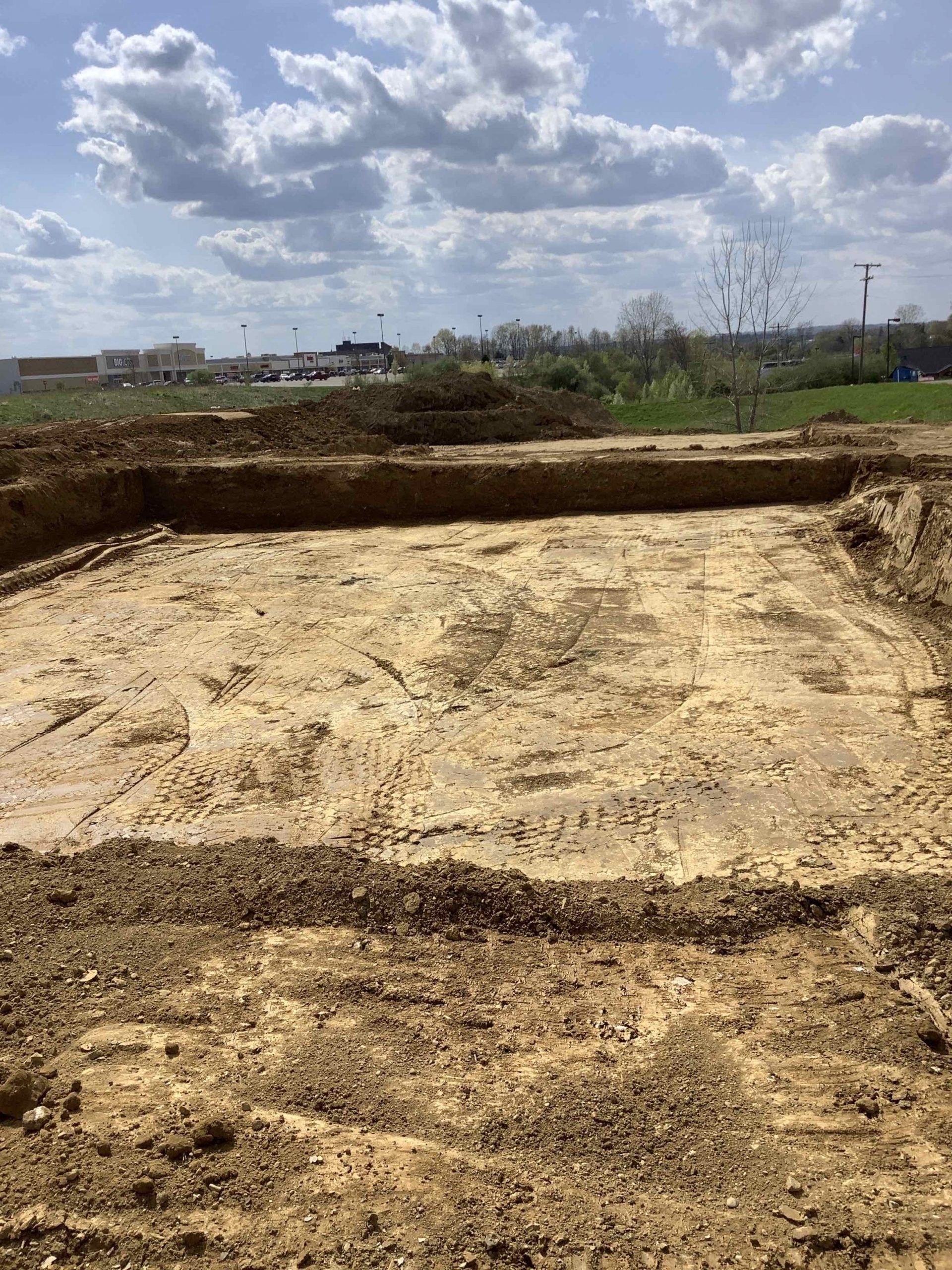 Excavated earth, square-shaped, under a cloudy sky. Dirt and mud visible, with distant buildings in the background.