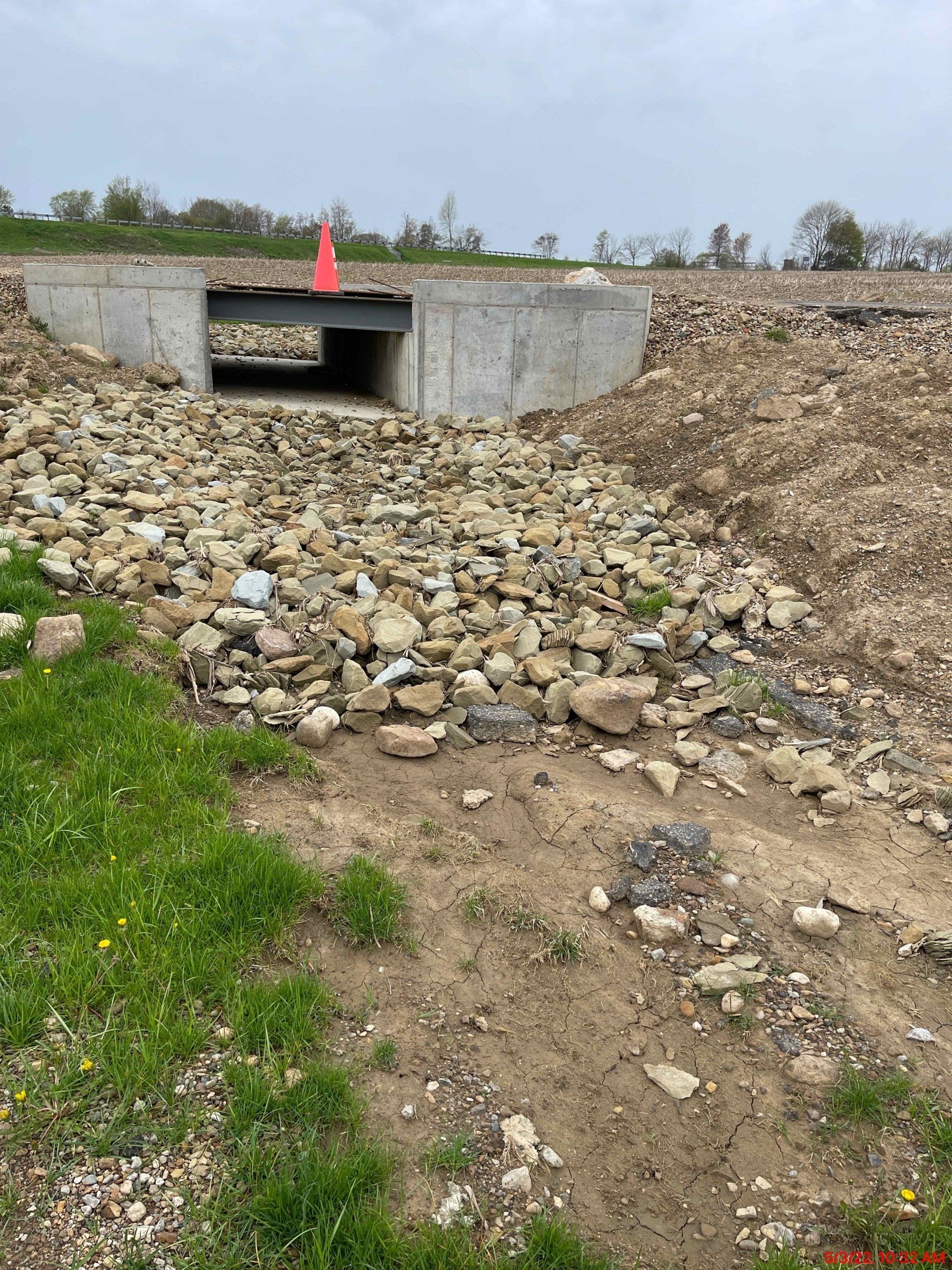 Concrete culvert under a road with a rocky streambed and a safety cone.
