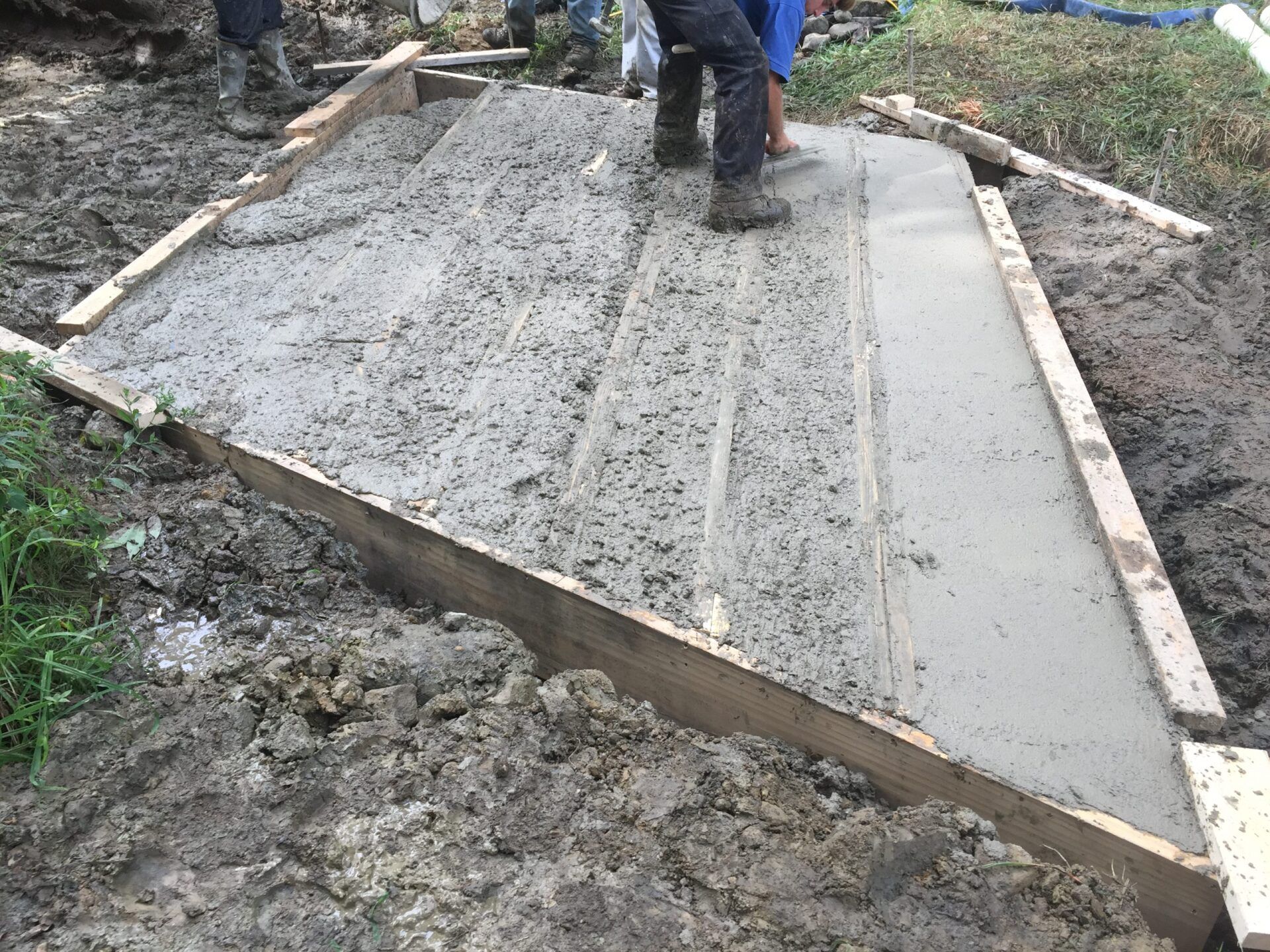 Fresh concrete poured within wooden forms, workers nearby in muddy surroundings.