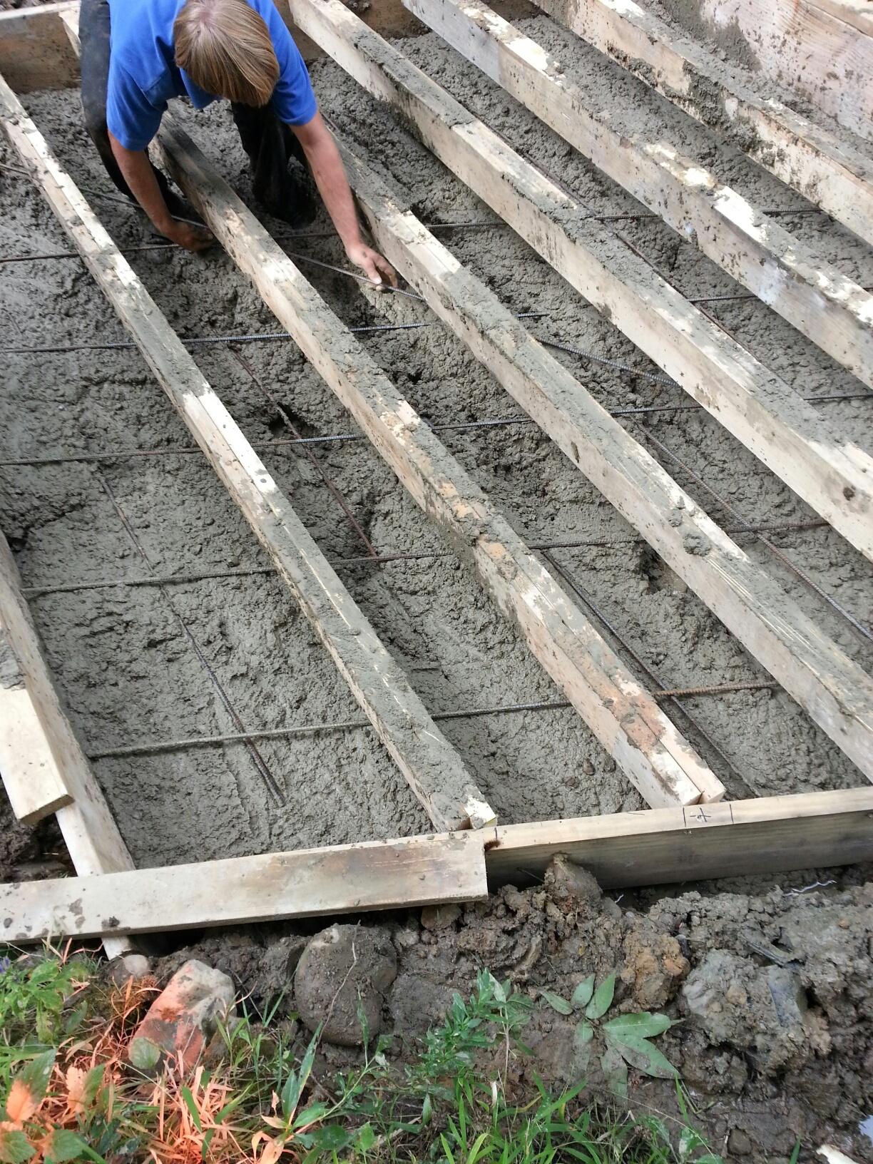 Person laying boards over wet concrete. Boards are parallel and within a wooden frame.