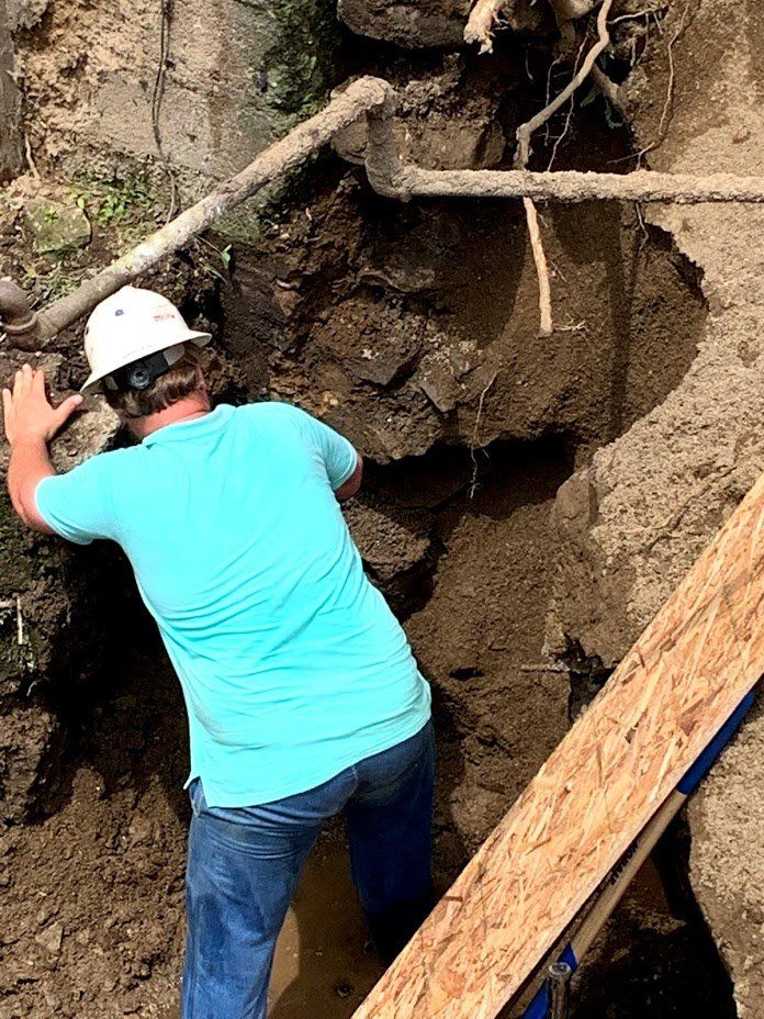 A person in a hard hat and blue shirt, working in a trench, examining a pipe.