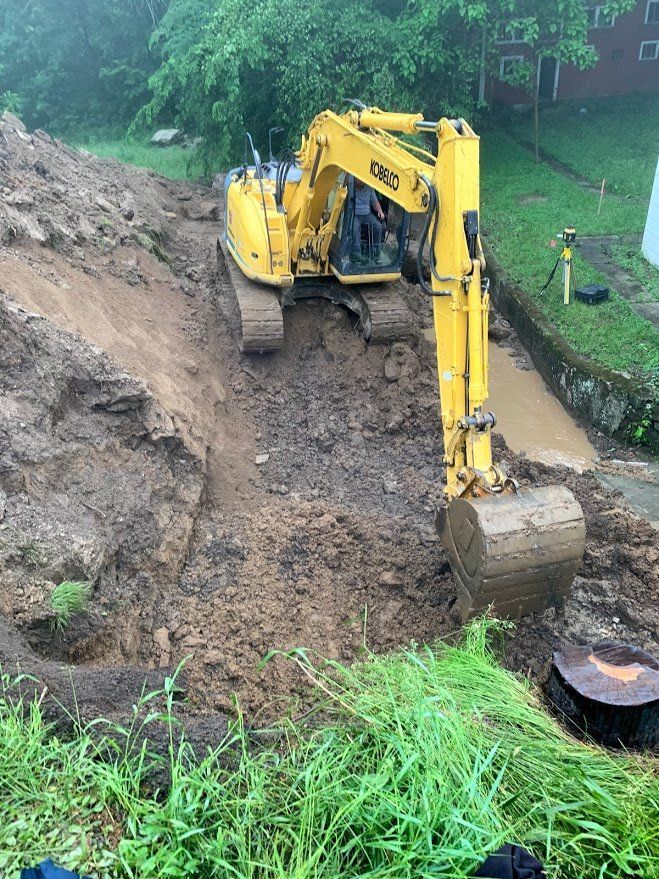 Yellow excavator digging in muddy soil next to a paved area and green vegetation.