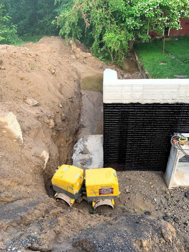 Construction site: trench next to concrete wall with black membrane, two yellow vibratory plate compactors.