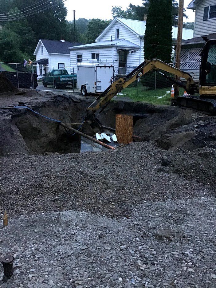 An excavator digs in a gravel-covered area. Houses and a trailer are in the background.