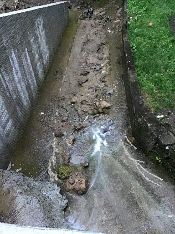 Muddy concrete drainage channel next to a wall and grass, with debris and water.