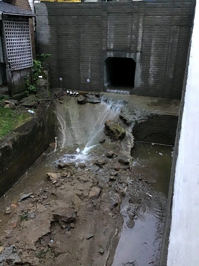 Drainage tunnel with muddy water spilling into an excavated area. Brick wall backdrop.