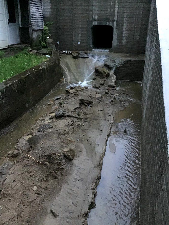 Muddy drainage channel with water flowing through a dark opening in a brick wall.