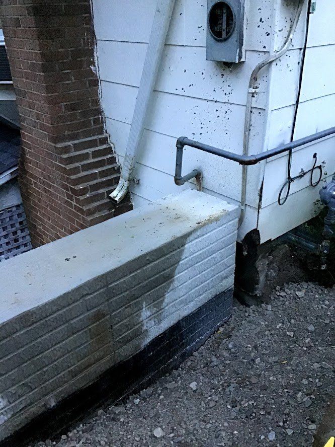 Exterior view: concrete wall against a building with electrical box, black pipe, and gravel ground.