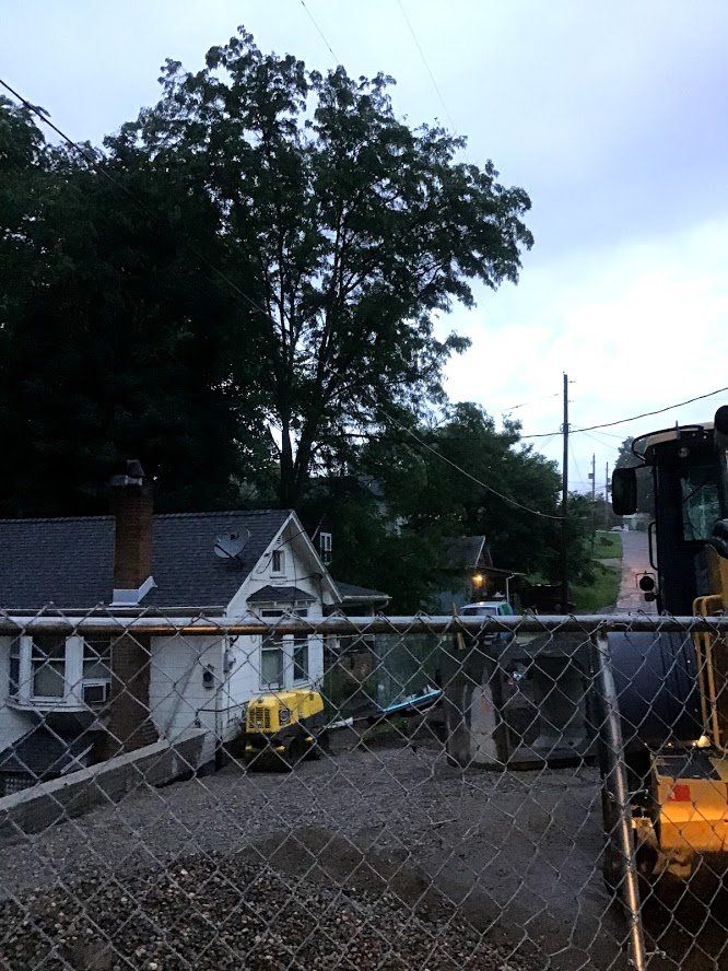 Chain-link fence in front of a white house, two yellow construction machines, trees, and power lines. Cloudy sky.