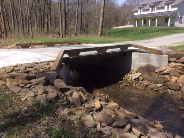 Concrete bridge over a creek in a rural setting; a large house is visible in the background.