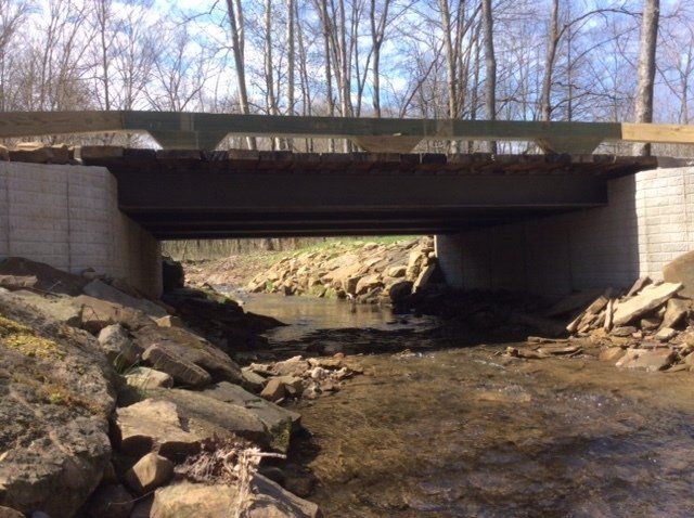 Bridge over a stream; brown wooden deck, concrete abutments, rocks in the water.