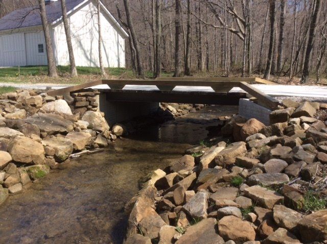 Wooden bridge over a creek, bordered by rocks, with a white building and trees in the background.