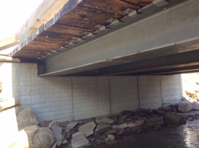 View from below a bridge with a textured concrete support wall and rocky riverbed.