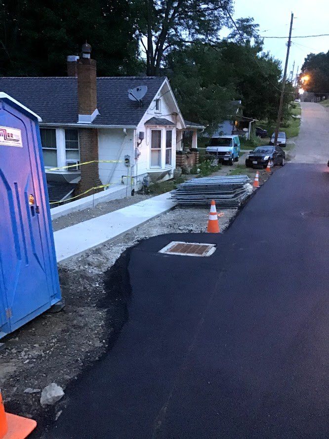 Street construction with new asphalt and sidewalk next to a house, porta-potty, and construction materials.
