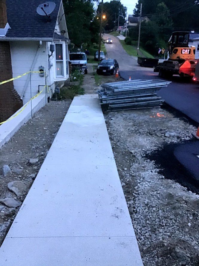 Sidewalk construction next to a house and street. Asphalt paving and heavy machinery visible.