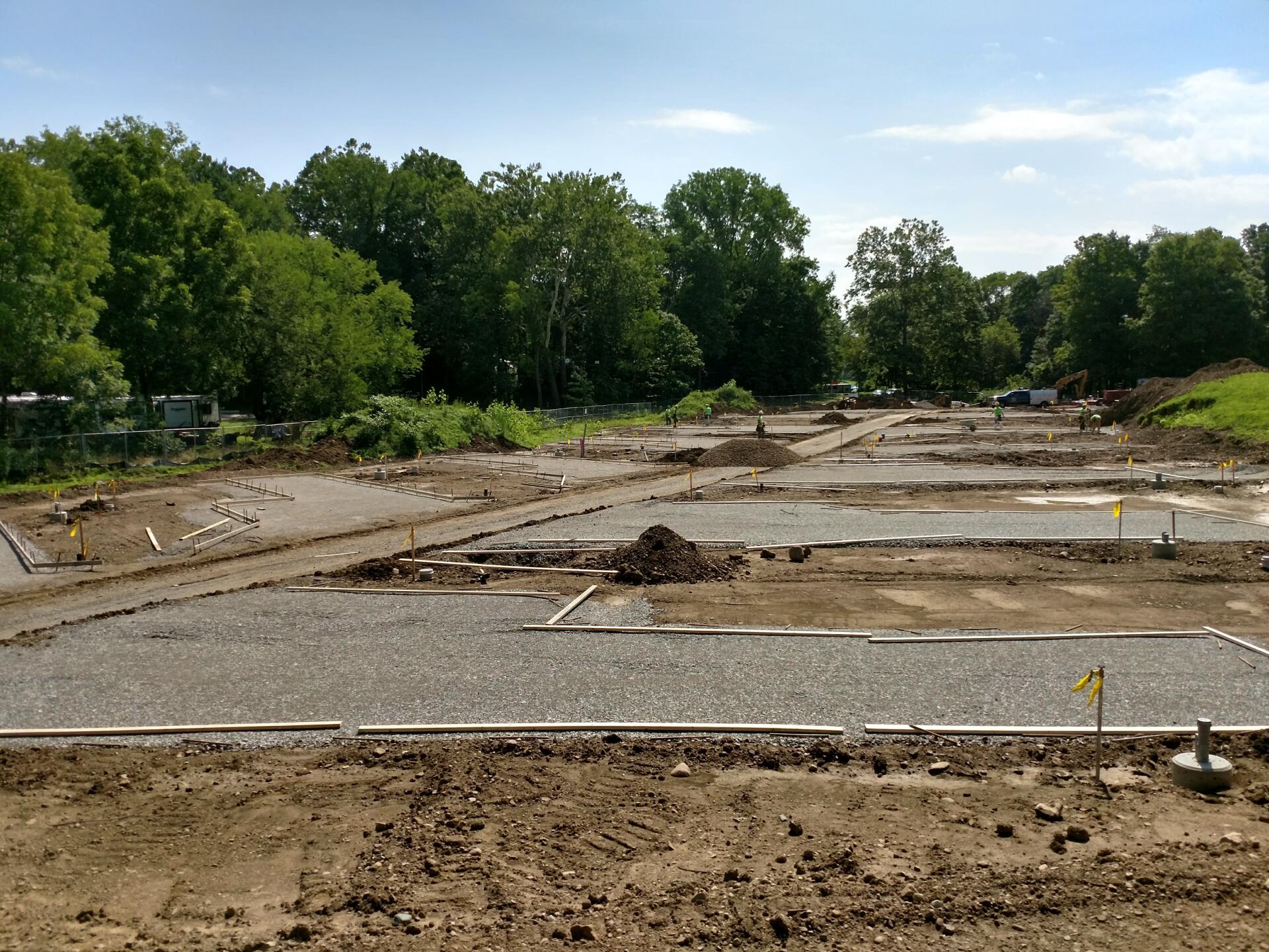 Construction site with gravel foundations, dirt, and trees in background under a blue sky.
