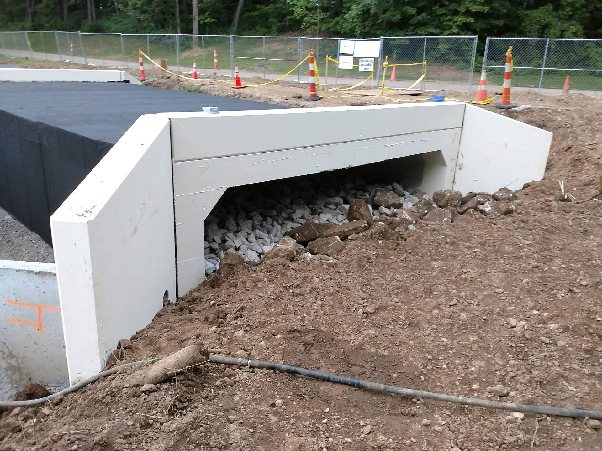 Concrete culvert under construction, surrounded by dirt and gravel. Black tarp on top, safety cones in the background.