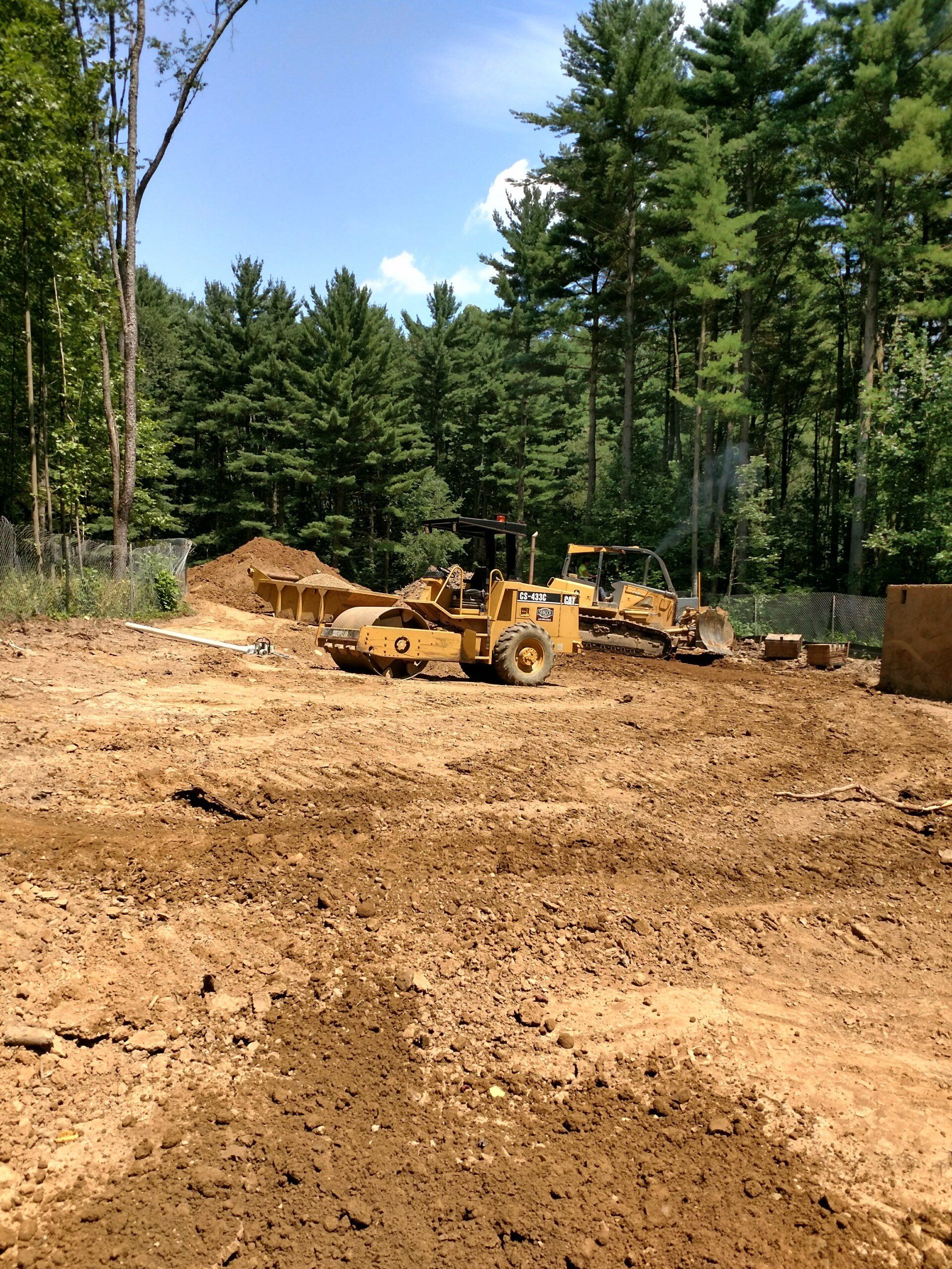 Construction site with earth-moving equipment, cleared ground, and a background of green trees under a blue sky.