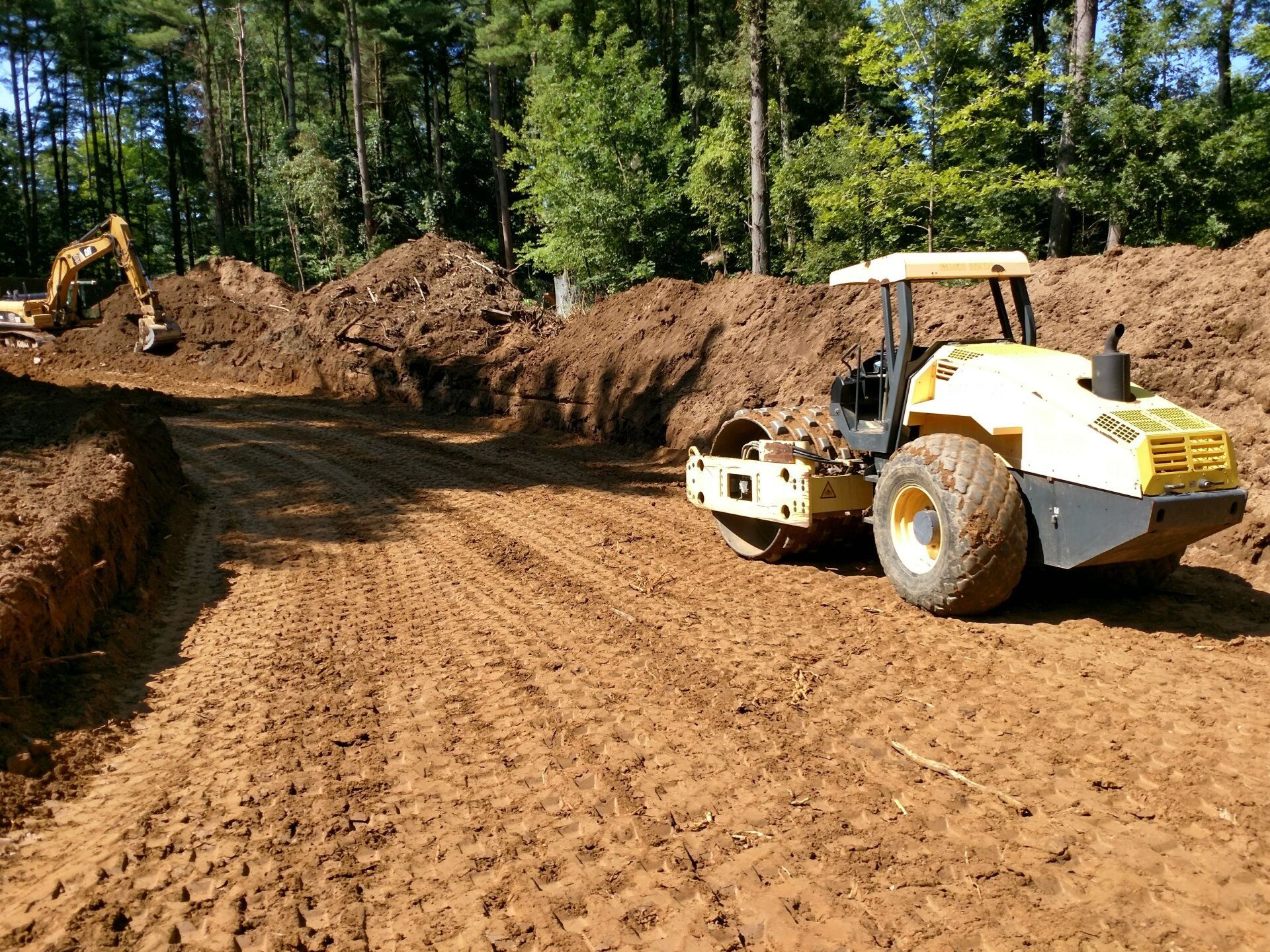 A yellow soil compactor levels a dirt road in a construction site with an excavator in the background.