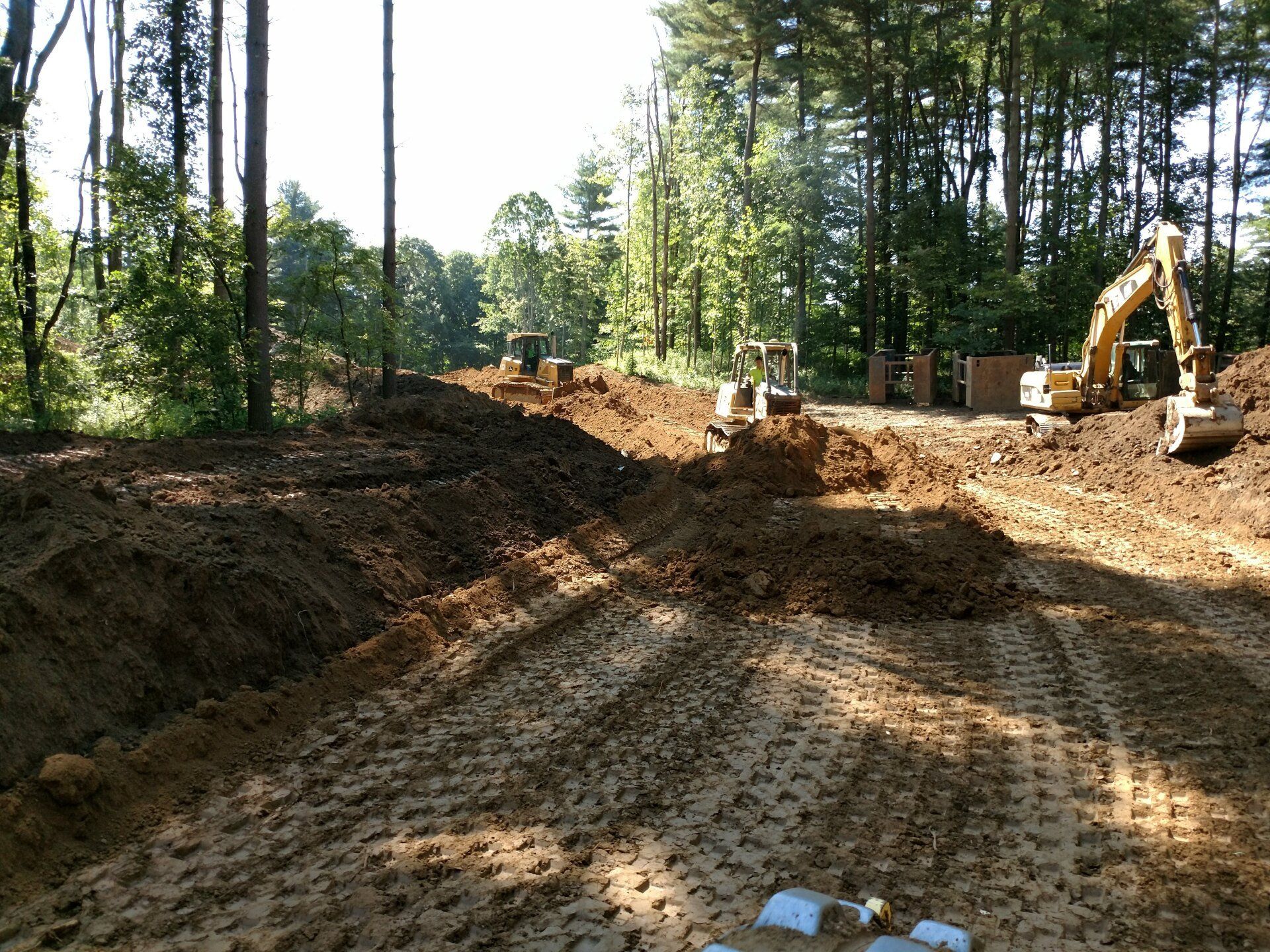 Construction site in a wooded area with heavy machinery, earth mounds, and sunlight.