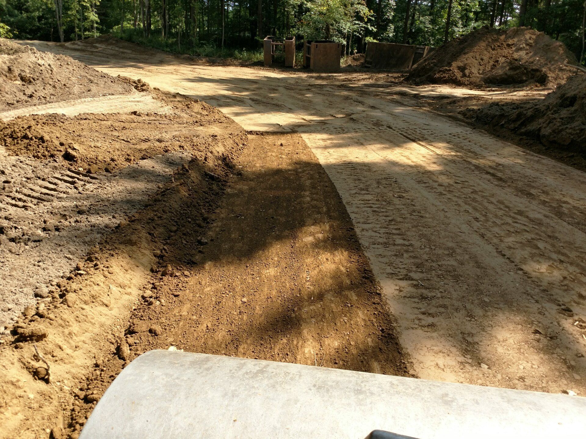 Dirt road under construction with tire tracks and piles of earth, concrete pipe in foreground.