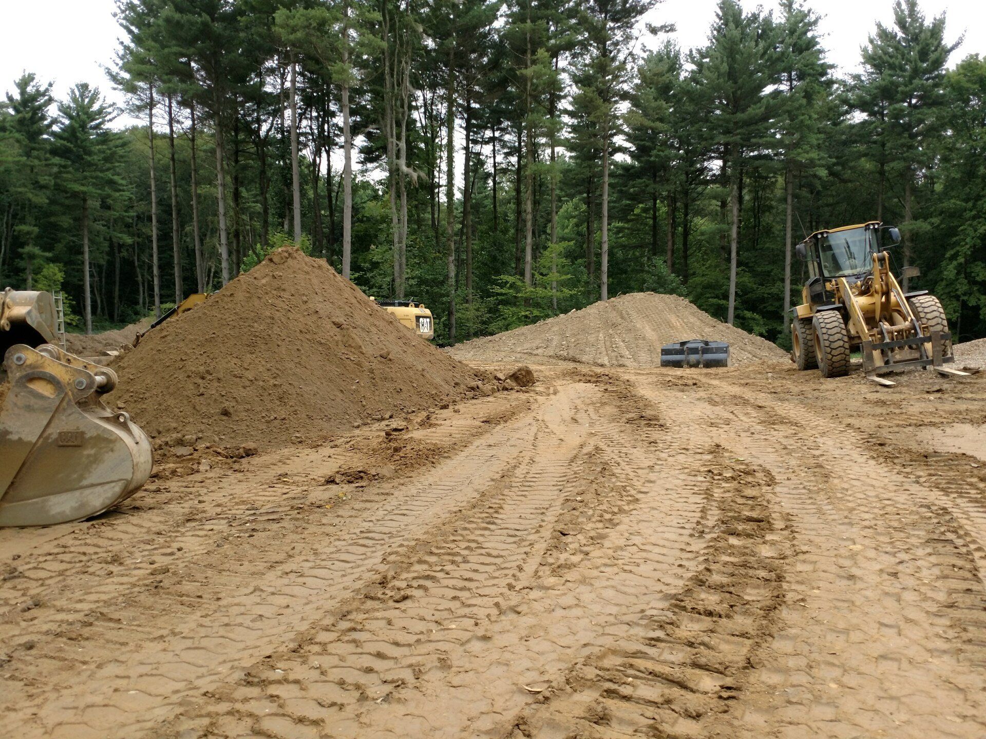 Construction site with piles of dirt and a yellow bulldozer, tire tracks, trees in background.