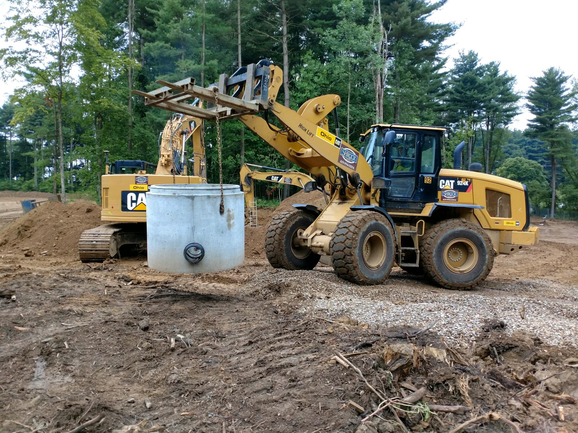 A yellow wheel loader and excavator lifting a concrete cylinder on a construction site.