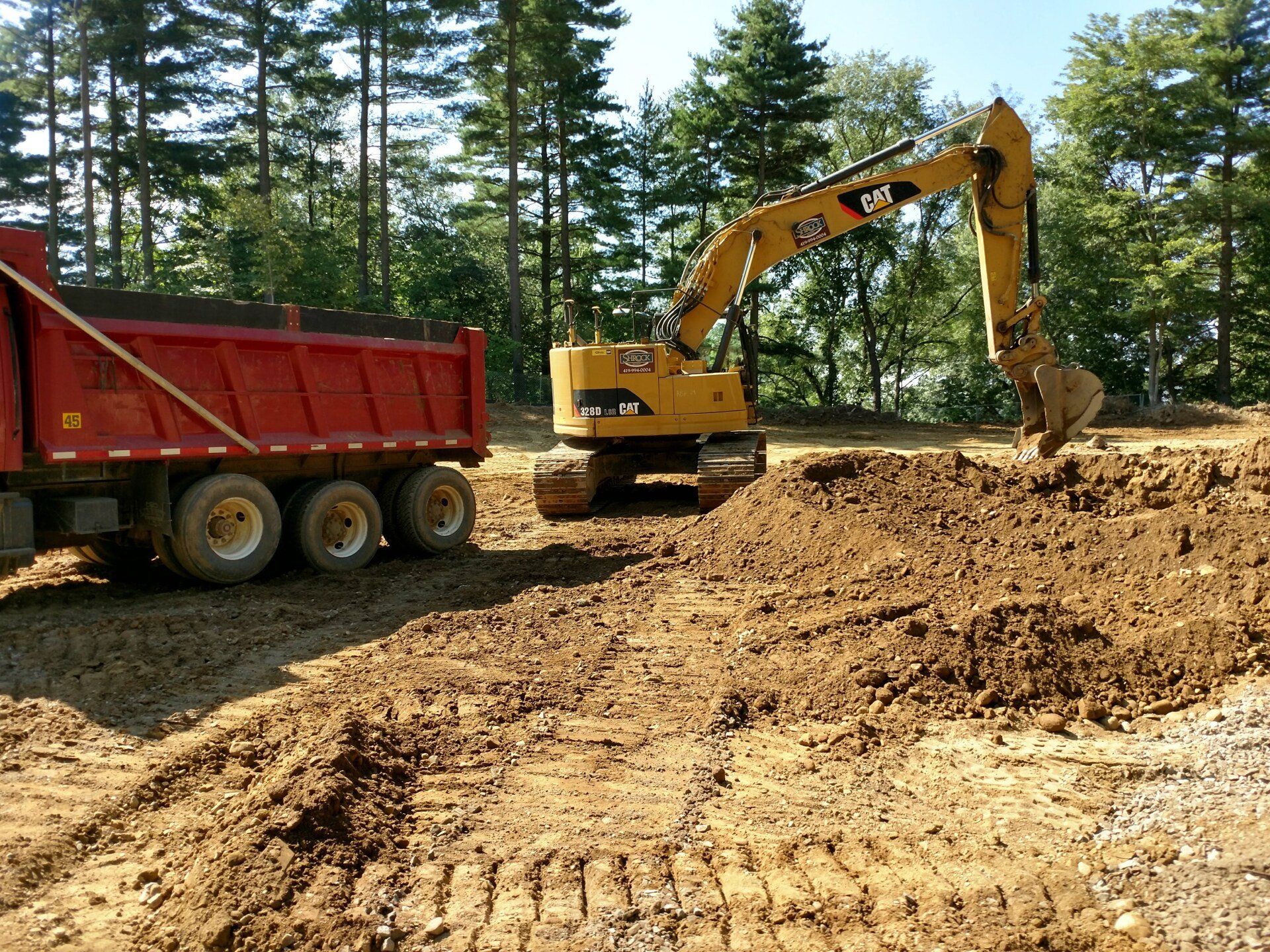 An excavator loading a red dump truck with dirt at a construction site in a wooded area.